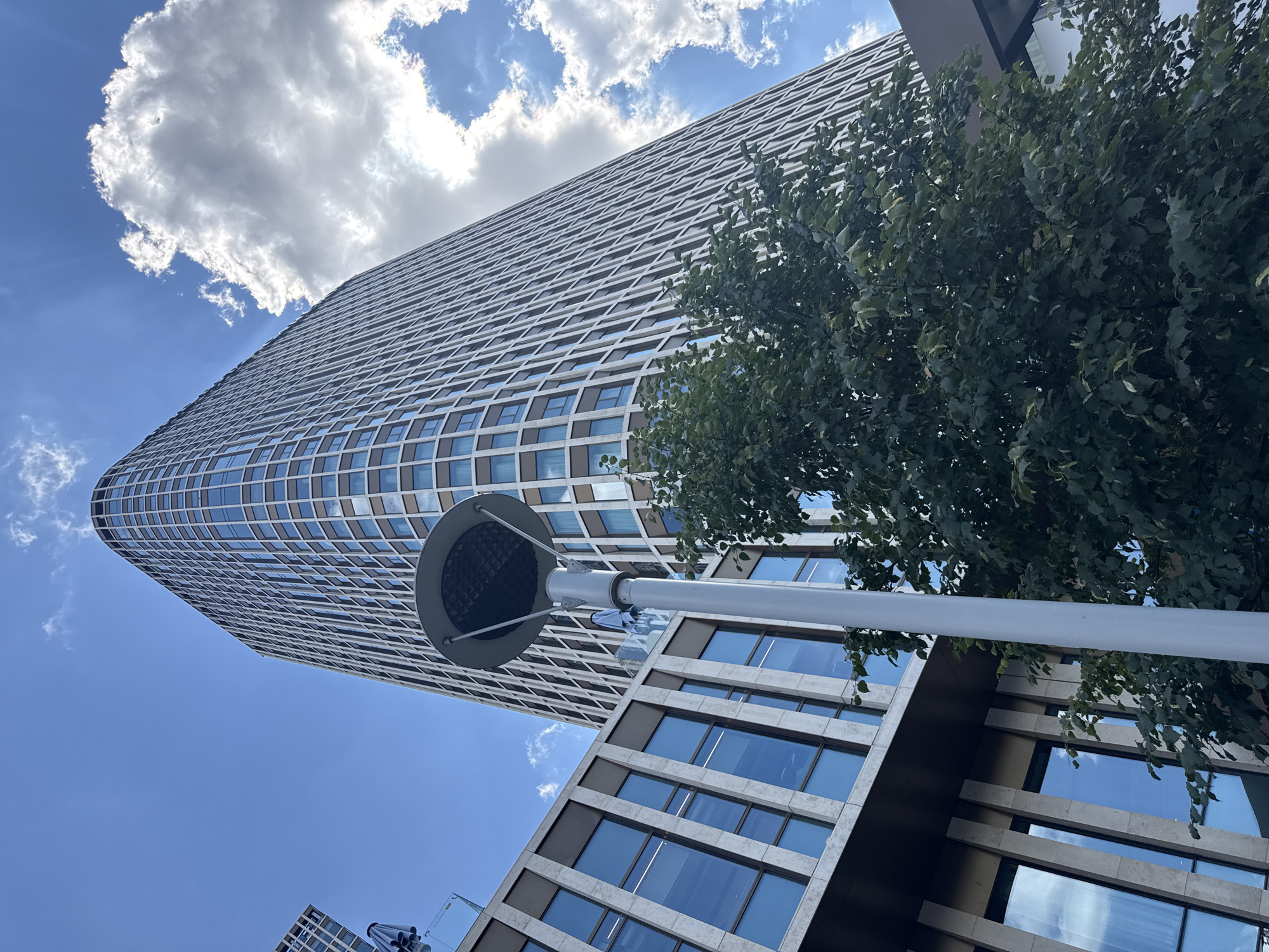 A low-angle, full shot captures a modern, curved skyscraper in Astana, Kazakhstan, dominating the frame against a vibrant blue sky dotted with fluffy white clouds. The skyscraper's facade is a grid of windows with beige frames, suggesting a sleek, contemporary design. To the left, a lower, rectangular building with similar architectural elements – large blue windows and beige vertical accents – is partially visible. A tall, white lamppost with a circular head stands in the foreground, slightly off-center, adding a vertical element to the composition. Lush green foliage from a tree obscures part of the lower building and extends upwards, softening the otherwise rigid lines of the architecture. The scene is bathed in natural daylight, indicating it was taken during the daytime.