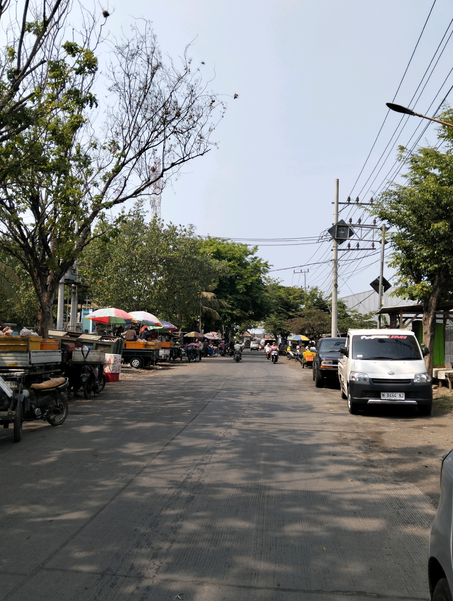 This is a daytime, eye-level, full-shot photograph of a street scene in Probolinggo, Indonesia. The street appears to be a busy local market area. On the left side of the road, there are several trucks with open beds, some carrying goods, and parked motorcycles. Colorful umbrellas are set up over some of the stalls. Several people are visible, some standing around the stalls and others on motorcycles. On the right side of the road, a white van with license plate "N 8464 NJ" is parked, and further down the road, other vehicles, including a black SUV, are seen driving or parked. Power lines and a street light are prominent in the background, with lush green trees lining the sides of the road and a tall communication tower visible in the distance. The sky is clear and bright.