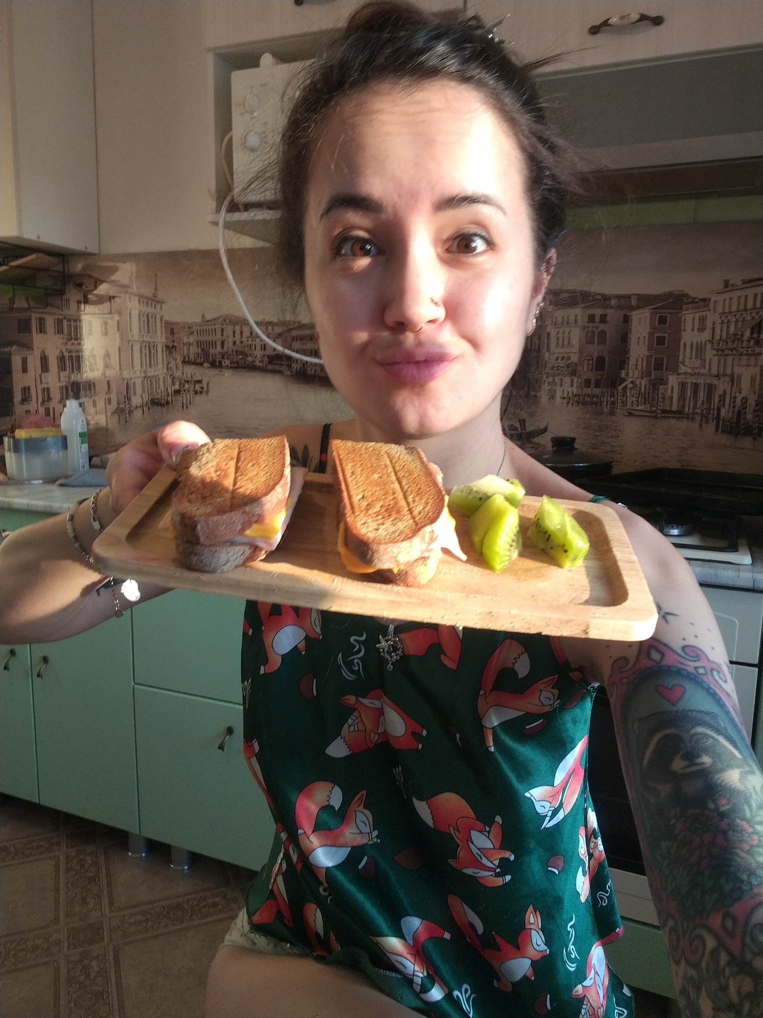 A woman is holding a wooden tray containing two sandwiches and cut kiwis. She is indoors, likely in a kitchen, with cabinets and a countertop visible in the background. The wallpaper behind her features a Venetian canal scene. She has dark hair pulled back, fair skin, and a nose piercing. She is wearing a green shirt with a fox pattern and has a tattoo on her left arm.