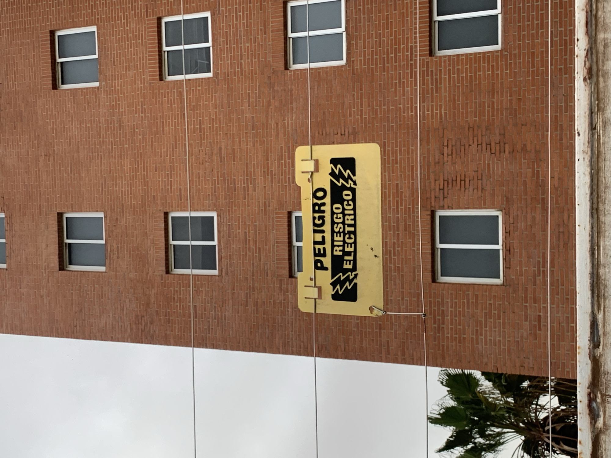 The media file displays an outdoor scene, captured and presented in a rotated orientation, appearing as if the camera was tilted 90 degrees counter-clockwise.
The primary subject is a multi-story brick building, which occupies the majority of the right side of the frame in its current orientation. The building features numerous rectangular windows with white frames and darkened panes, arranged in a grid-like pattern.
Hanging against the brick wall, near the center of the image, is a bright yellow rectangular sign. The sign is suspended by wires and features bold, black text and symbols. When the image is viewed correctly (rotated 90 degrees clockwise), the text reads "PELIGRO RIESGO ELECTRICO" along with a lightning bolt symbol, indicating "DANGER ELECTRICAL RISK" or "ELECTRIC HAZARD."
Multiple thin wires run vertically across the brick facade, some appearing to support the yellow sign, others possibly utility lines. To the far right, a weathered utility pole or structural column runs vertically along the edge of the frame.
The bottom left portion of the image (when rotated) shows an overcast sky, suggesting cloudy weather conditions during daytime. A portion of a green palm tree is visible in the very bottom right corner, partially obscured. The overall atmosphere is mundane and functional, with no human activity or strong emotions conveyed. The architecture and vegetation are consistent with the provided location context of Pampatar, Venezuela.