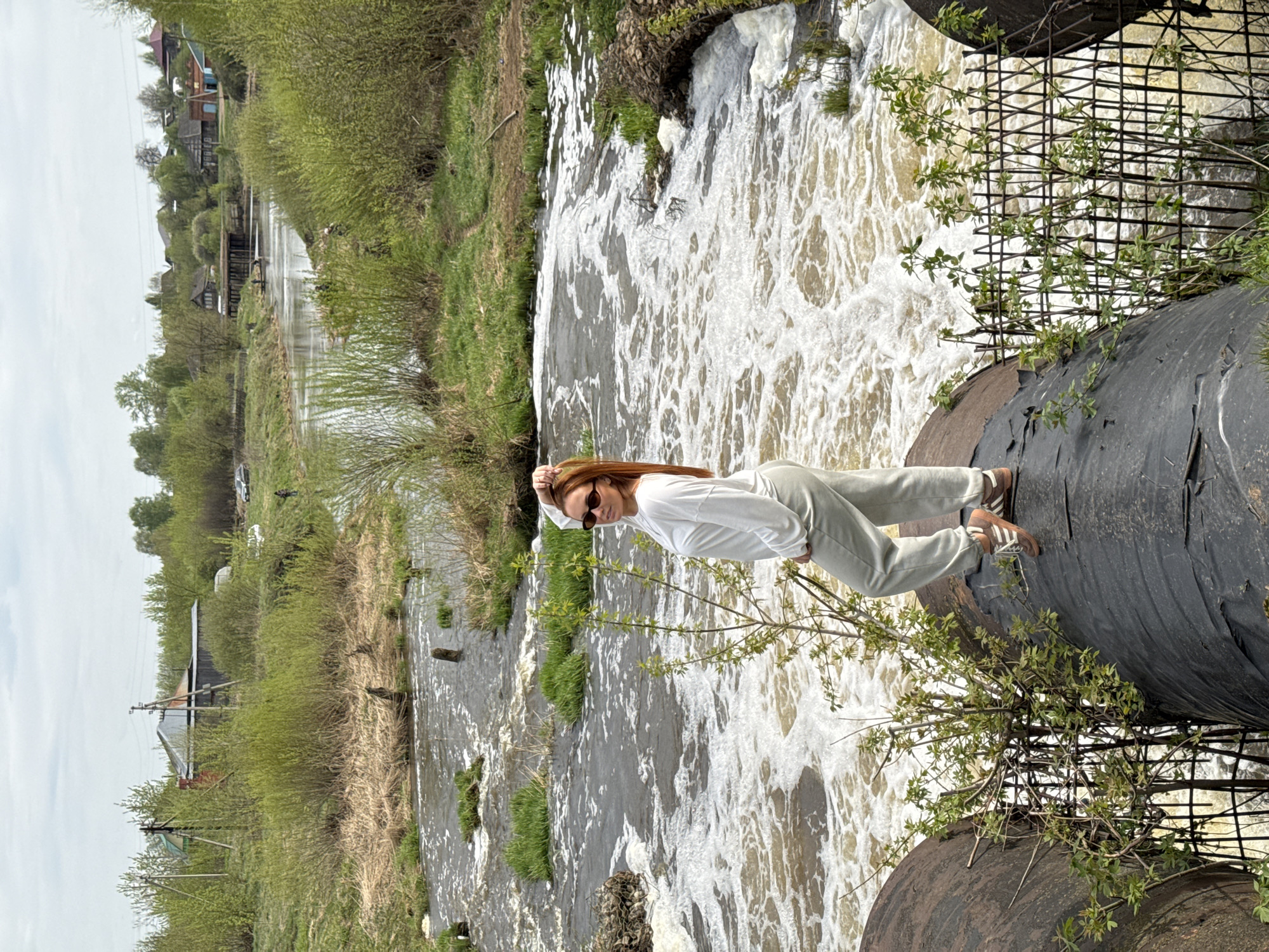 A woman stands on a large, black pipe, presumably a section of a water or sewage system, that spans a turbulent river or stream. The water is foamy and churns below her, indicative of a small waterfall or rapid. She is dressed in light-colored clothing and sunglasses. The environment around her consists of green vegetation, including trees and shrubs. In the background, structures resembling residential buildings can be seen, possibly houses or small buildings. The image is taken outdoors under daylight conditions. The location is Nozhovka, Russia.