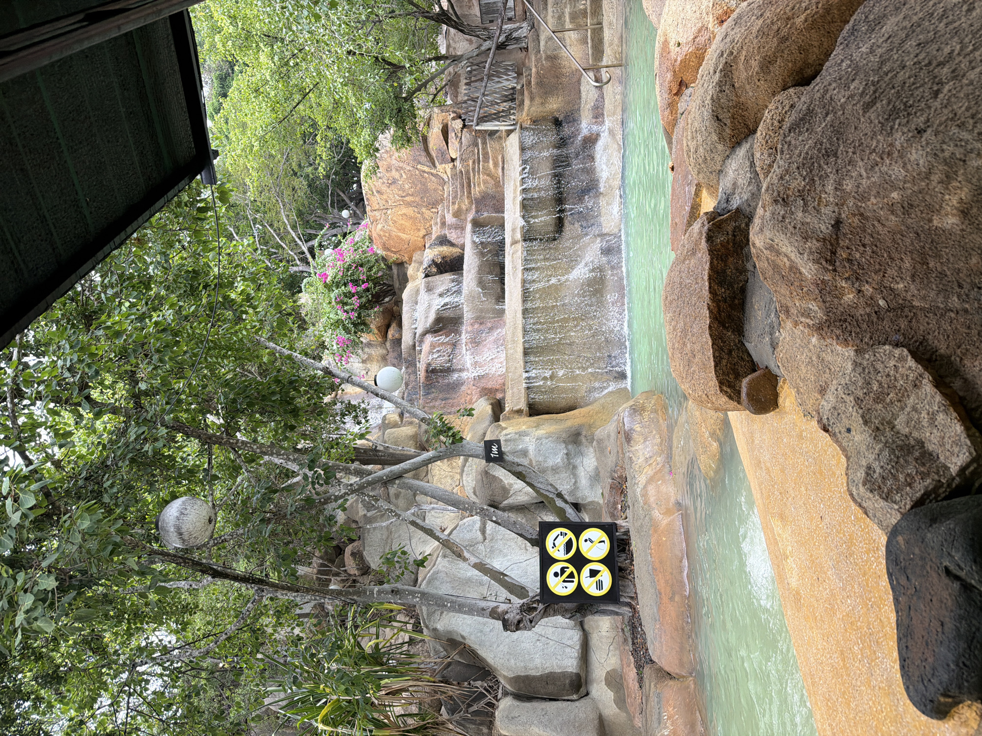 The image shows a man-made rock waterfall cascading into a shallow pool with turquoise water. The pool area is surrounded by large rocks and lush green vegetation. A sign with four prohibition symbols is posted on a tree near the waterfall, indicating rules such as no running, no diving, no eating or drinking, and no smoking. Another sign on a nearby tree reads "1M", likely indicating a depth of one meter. In the foreground, large rocks and a sandy embankment are visible. The scene appears to be a part of a resort or recreational area, possibly in Nha Trang, Vietnam, given the tropical foliage and the architecture of the covered area in the upper right corner.