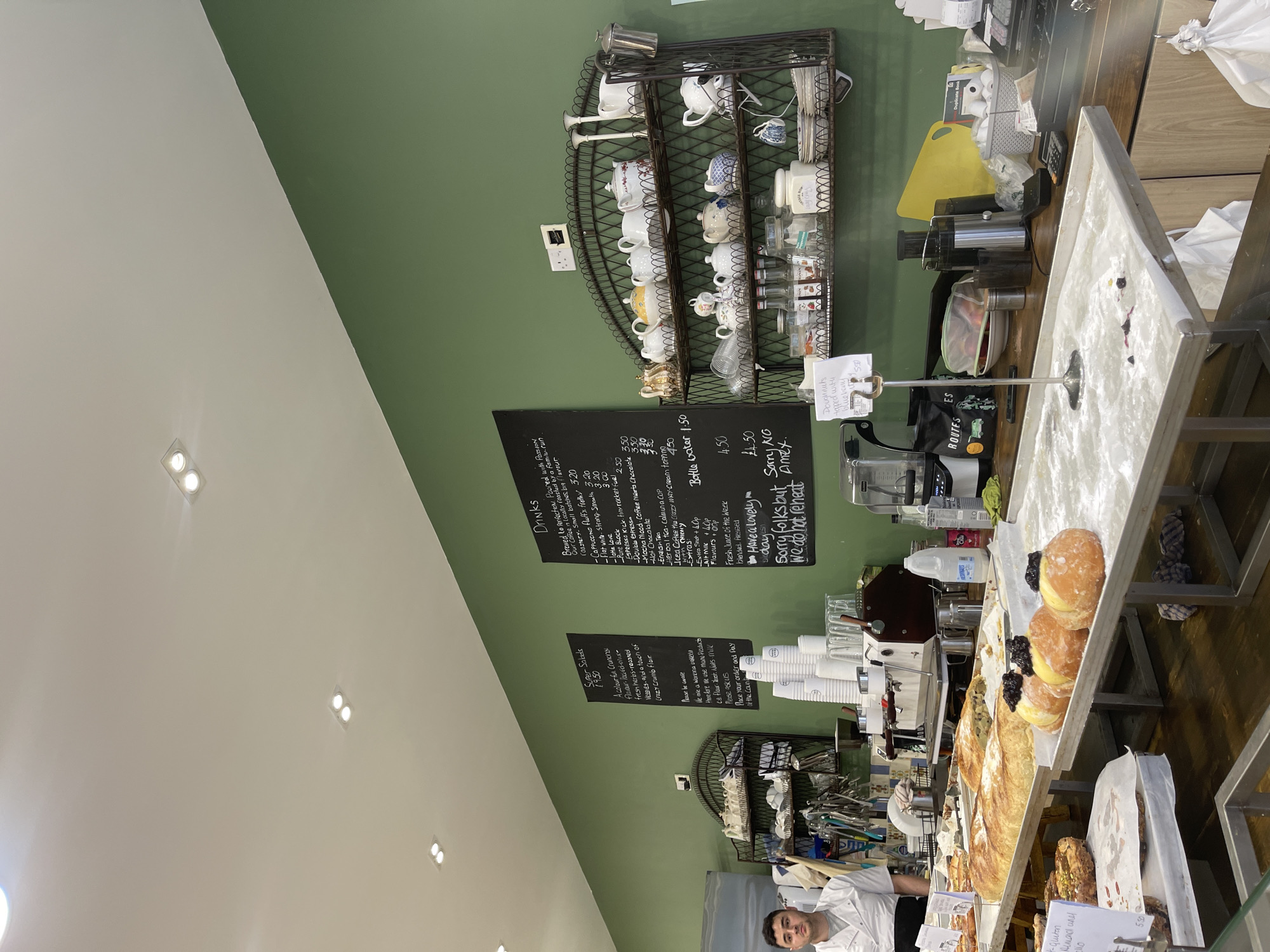 Here's a detailed description of the image:
**Setting:** The image appears to be inside a bakery or café. The walls are painted a matte green, and a white ceiling can be seen. The scene is likely set in Portsmouth, United Kingdom, based on the mention of "Our Coffee" and recognizable UK currency.
**People:** A person wearing a white chef's coat is partially visible at the bottom of the frame, likely a staff member behind the counter.
**Objects:**
* **Counter:** A counter displays various pastries, including doughnuts with what appears to be blueberry topping. There is also other baked goods such as a loaf of bread. A sign notes that there are gluten-free almond options.
* **Drinks Menu:** Two blackboards display drinks and super salads menus. The menu includes items like cappuccinos, lattes, mochas, herbal teas, and a "fresh juice of the week." Prices are listed in UK currency (£).
* **Coffee Station:** Behind the counter, there are espresso machines, blenders, and other equipment needed for preparing drinks. A rack holds teacups and other items.
* **Signage:** Signs at the till indicates "Sorry folks but same way we do not reheat". There is also a sign for payment methods indicating that they don't accept Amex.
**Actions and Interactions:**
* A staff member appears to be working behind the counter.
* The environment is set for serving and displaying baked goods and beverages to customers.
**Overall Impression:** The image captures a scene inside a bakery or café with a focus on its offerings, including pastries and drinks. The setup appears well-organized.