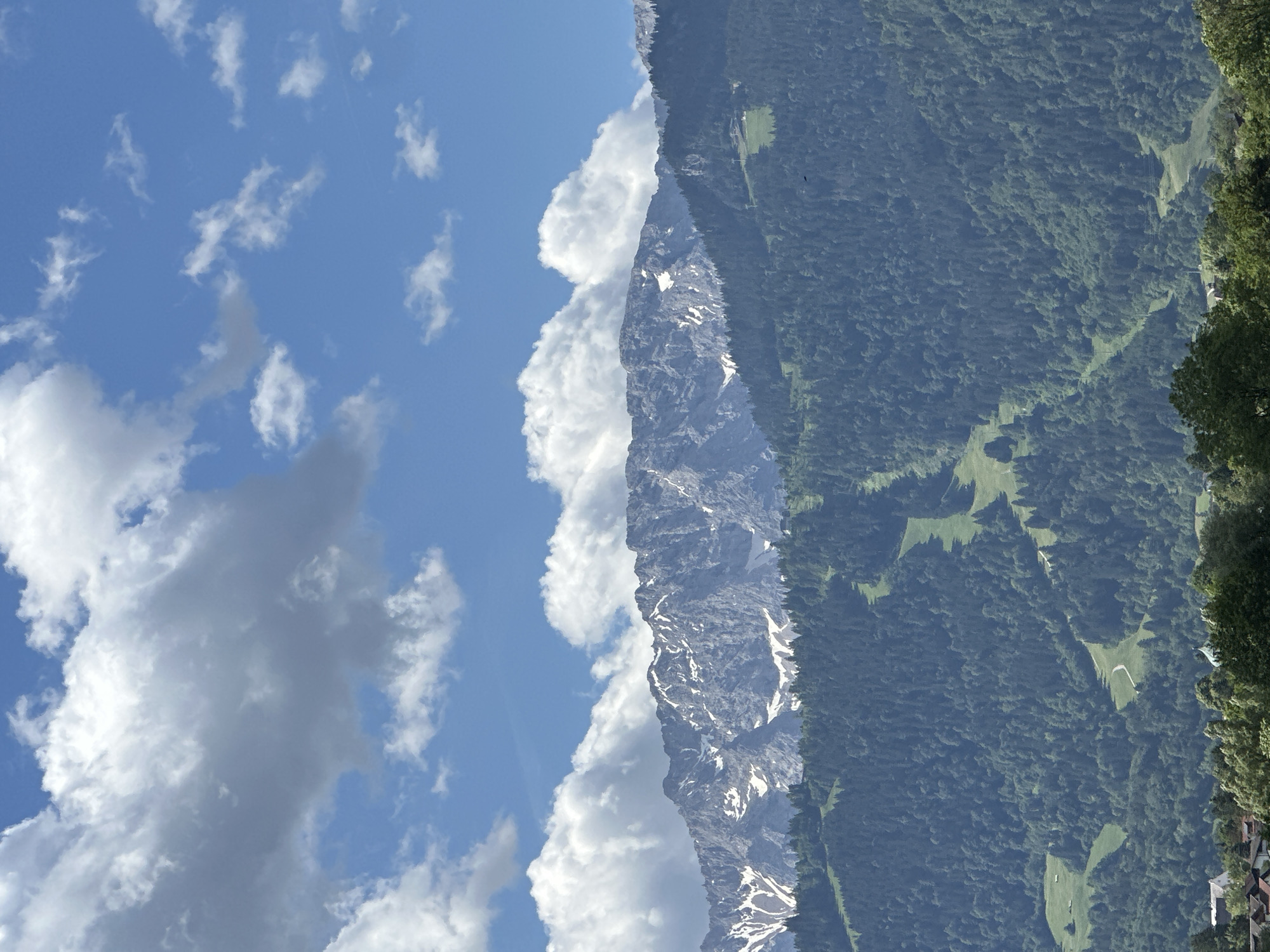 The image shows a view of the Alps from Garmisch-Partenkirchen, Germany. In the foreground, there is a dense forest of evergreen trees covering a mountainside. A winding path or ski slope cuts through the trees. In the mid-ground, a rugged mountain range with snow-capped peaks rises above the treeline. The sky above is a vibrant blue with scattered white cumulus clouds. The overall scene is one of natural beauty and tranquility, characteristic of the Bavarian Alps.