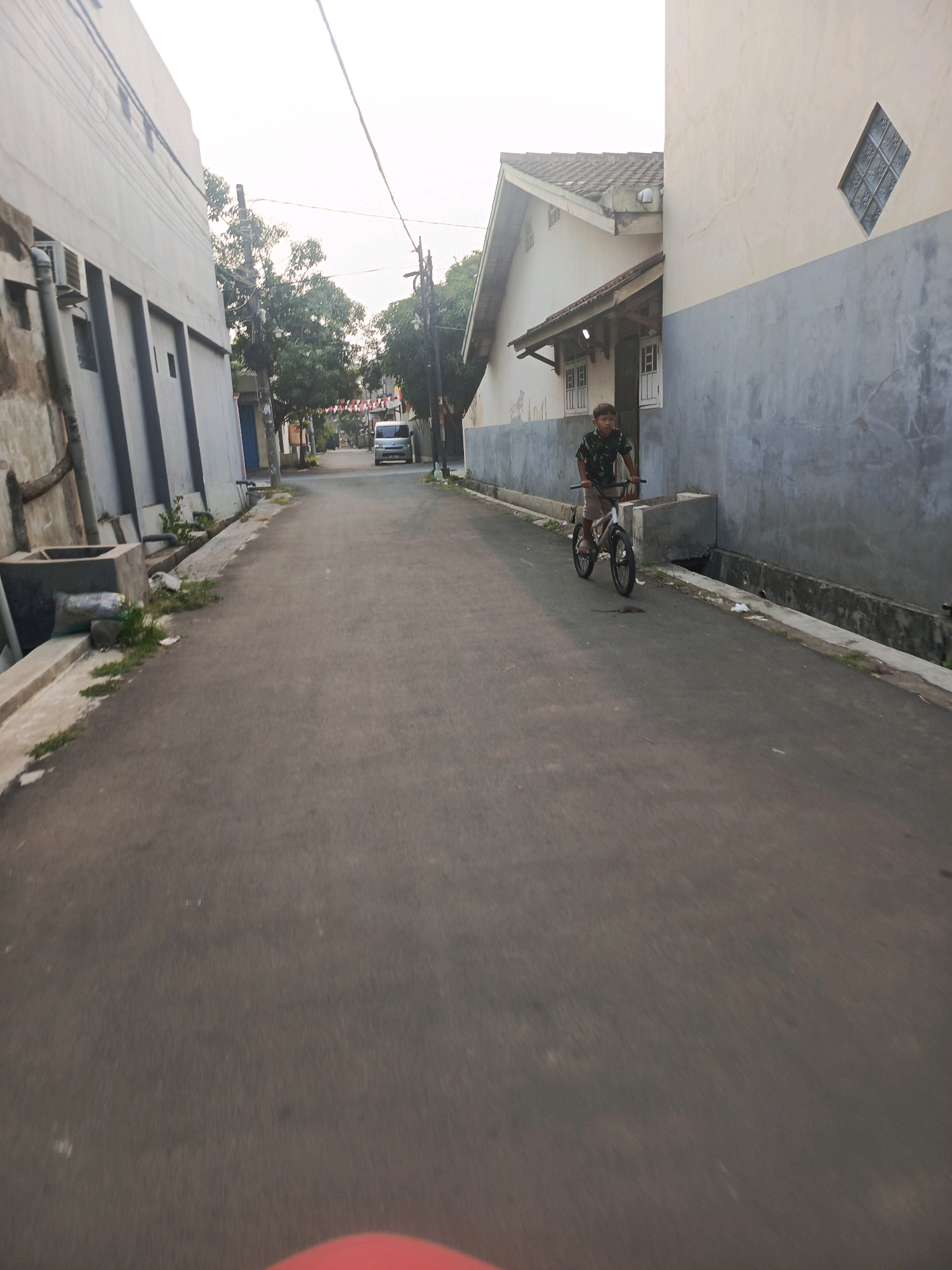 The image depicts a street scene in Balaraja, Indonesia, likely during the daytime due to the visible lighting conditions. In the foreground and midground, a young boy is riding a bicycle along an asphalt road. He is wearing a dark-colored patterned shirt and light-colored shorts. The bicycle appears to be a BMX-style bike.
The environment consists of a narrow street flanked by buildings on both sides. On the left, a multi-story building with light-colored walls and dark vertical accents is visible, featuring an external pipe system and some concrete structures at its base. On the right, a two-story building with a light-colored upper facade and a darker, textured lower facade is prominent. This building has a pitched roof with tiles and a diamond-shaped window made of glass blocks.
Further down the street, a silver van or small SUV is parked. The street is lined with some greenery and what appear to be utility poles with wires. Decorative red and white flags are strung across the street in the distance, suggesting a festive occasion or holiday. The road surface shows some wear and tear. The sky is overcast or hazy, contributing to diffused lighting.