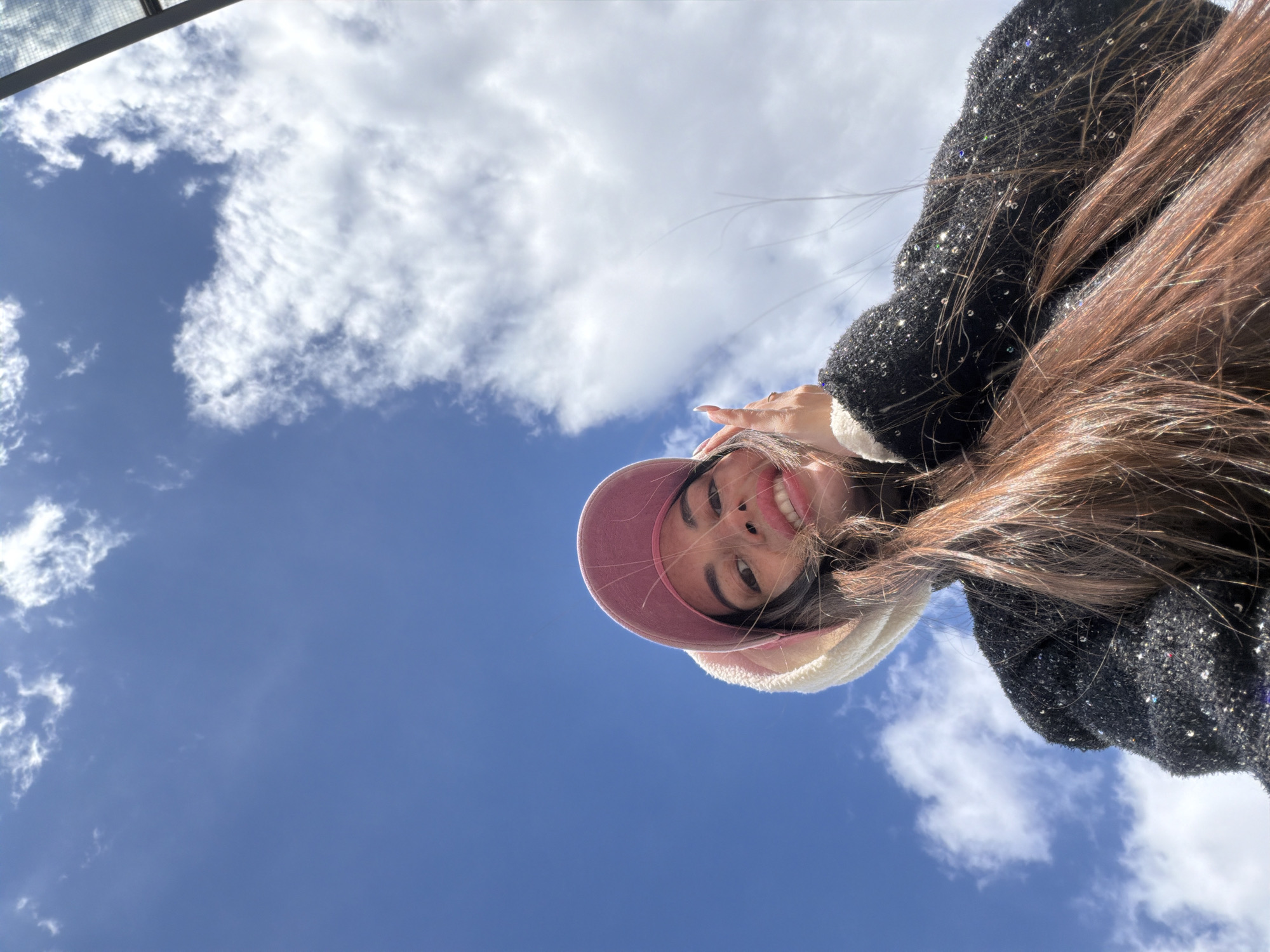 A vibrant, low-angle selfie captures a young woman smiling broadly and looking down towards the camera, set against a bright blue sky dotted with fluffy white clouds. The image conveys a cheerful and pleasant mood, likely taken outdoors during daytime in Saint Petersburg, Russia.
The subject, a young woman, is positioned in the upper portion of the frame, appearing inverted relative to the ground. She wears a pink visor or cap, layered over a white, fluffy hat or ear muffs. Her long, brown hair flows around her face and shoulders. Her attire includes a dark-colored garment, possibly a jacket or top, adorned with small, sparkling sequins that reflect the sunlight, adding a shimmering detail to her appearance.
The background is dominated by a clear, azure sky with scattered cumulus clouds, indicating fair weather. The perspective is from directly below the subject, creating an upward-looking shot where the sky fills most of the frame. A subtle, metallic grid-like structure is partially visible in the extreme top-left corner, suggesting an architectural element overhead, such as a skylight or a structural beam. No text is visible in the image.