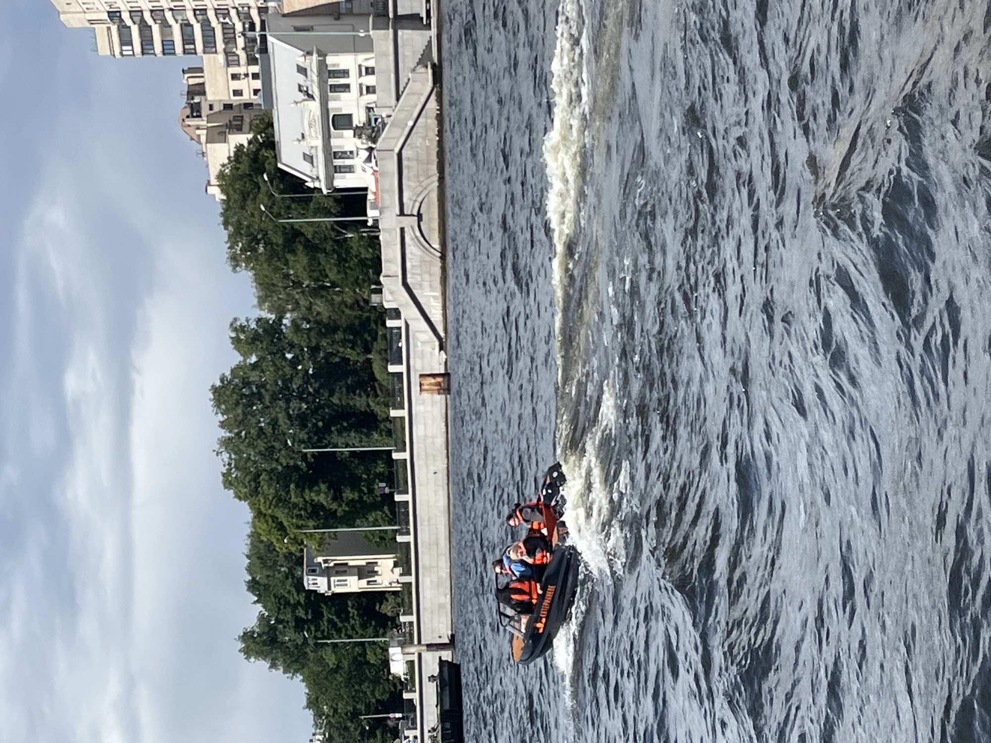The image depicts a scene in Saint Petersburg, Russia, likely during the daytime given the bright sky and clear visibility. The photo is oriented vertically, showing a body of water with a small, black motorboat cutting through the waves. The boat carries at least three individuals wearing life vests. One person appears to be at the helm, while others are seated. The boat has text on its side, but it is difficult to read clearly.
In the background, a waterfront promenade or embankment is visible, constructed with stone steps and railings. Beyond the embankment, lush green trees line the shore, suggesting a park or a tree-lined district. Further in the distance, several multi-story buildings, characteristic of urban architecture, are present, hinting at a city setting. The sky is partly cloudy, with white clouds against a light blue background. The water is choppy, with white foam trailing behind the boat, indicating it is moving at a moderate speed. The overall impression is that of an active recreational outing or possibly a water rescue demonstration on a pleasant day in Saint Petersburg.