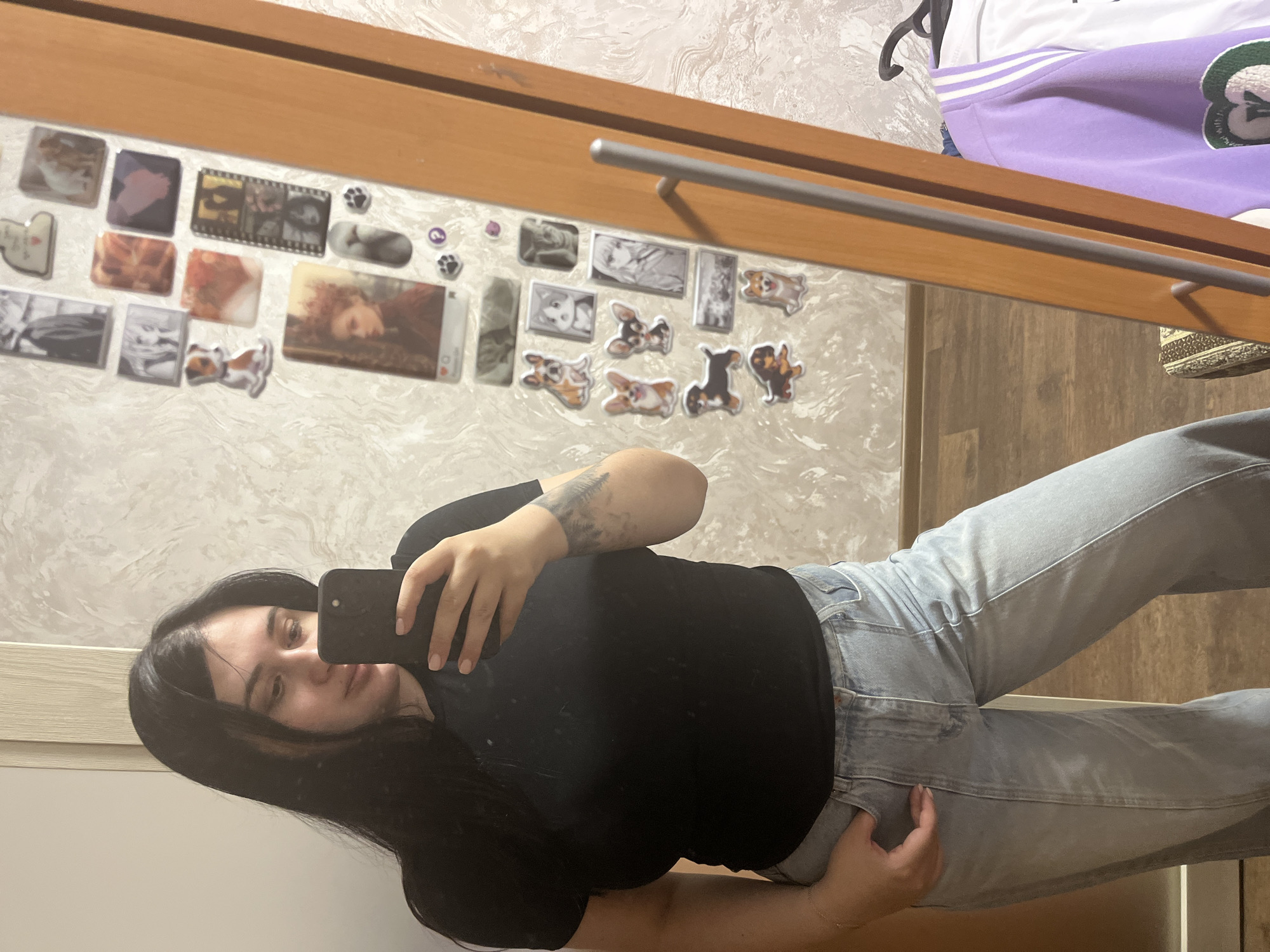 A young woman is depicted taking a mirror selfie indoors in Chaikovsky, Russia. She has long, dark hair and is dressed in a black t-shirt and light blue jeans. Her left arm features a tattoo, and she is holding a black smartphone to capture her reflection. Her facial expression appears neutral.
The mirror is framed with light-colored wood, and a horizontal silver metal rod is attached to the frame, possibly for hanging items. The wall behind the mirror is covered in a light-toned, textured wallpaper.
The mirror's surface is extensively decorated with various stickers or magnets. These include numerous images of cartoon or stylized dogs, particularly Corgis, alongside some cat pictures. Other notable decorations include a black and white film strip, a sticker resembling an Instagram post with a "like" heart icon, and one that reads "I HEART U". A small round sticker shows an 'R' (or 'Я' in Cyrillic) and a '2'. In the upper right corner of the reflection, a purple and white garment is visible hanging, suggesting a casual home environment. The floor visible in the reflection is made of wood.