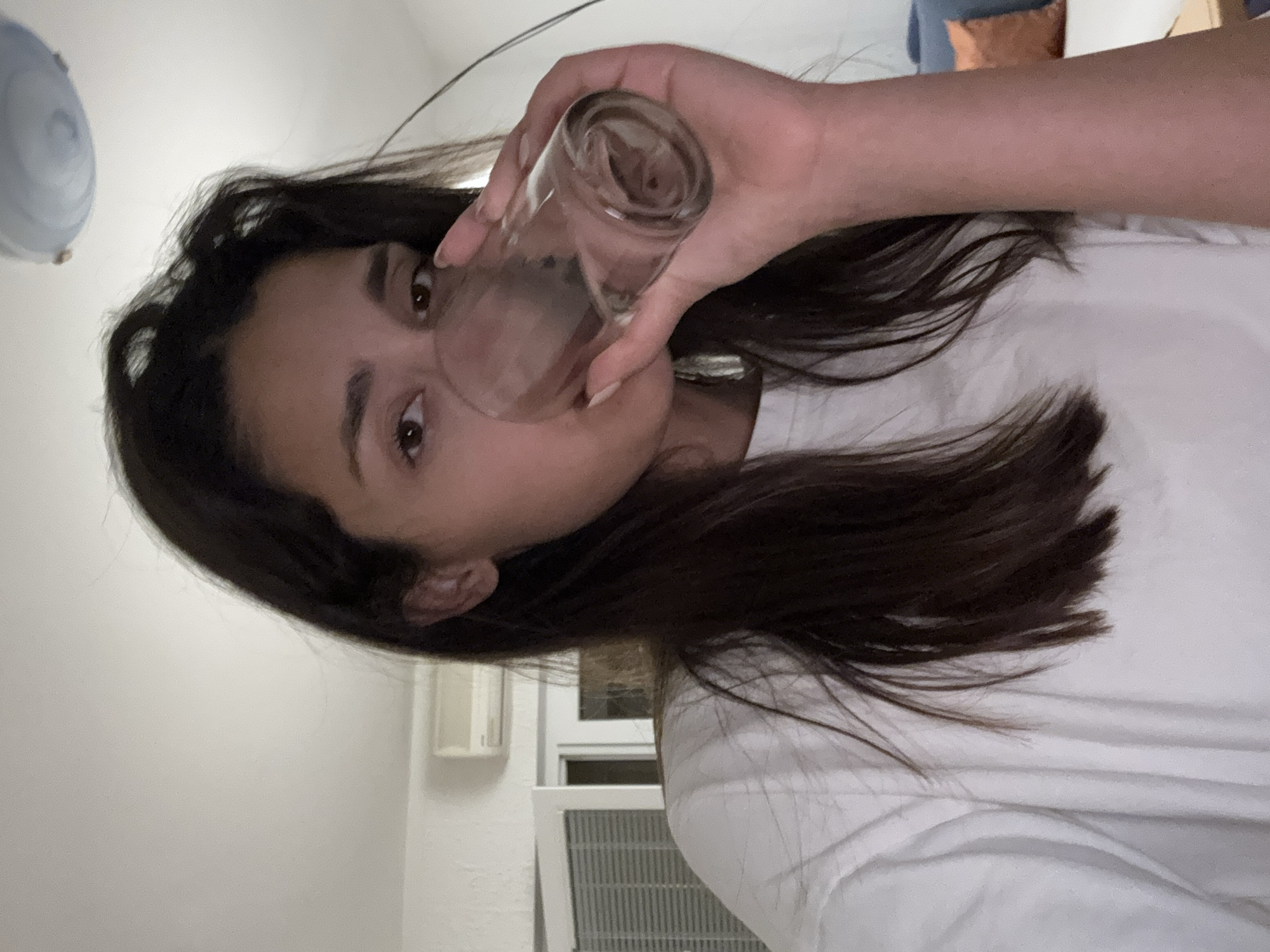 A young woman is drinking from a clear glass. She has dark brown hair and is wearing a white t-shirt. The background includes a white wall, an air conditioning unit, and a glimpse of a blue cushion or couch. The image appears to be taken indoors. There is no visible evidence in this photograph indicating a location in the city of Sutomore, Montenegro.