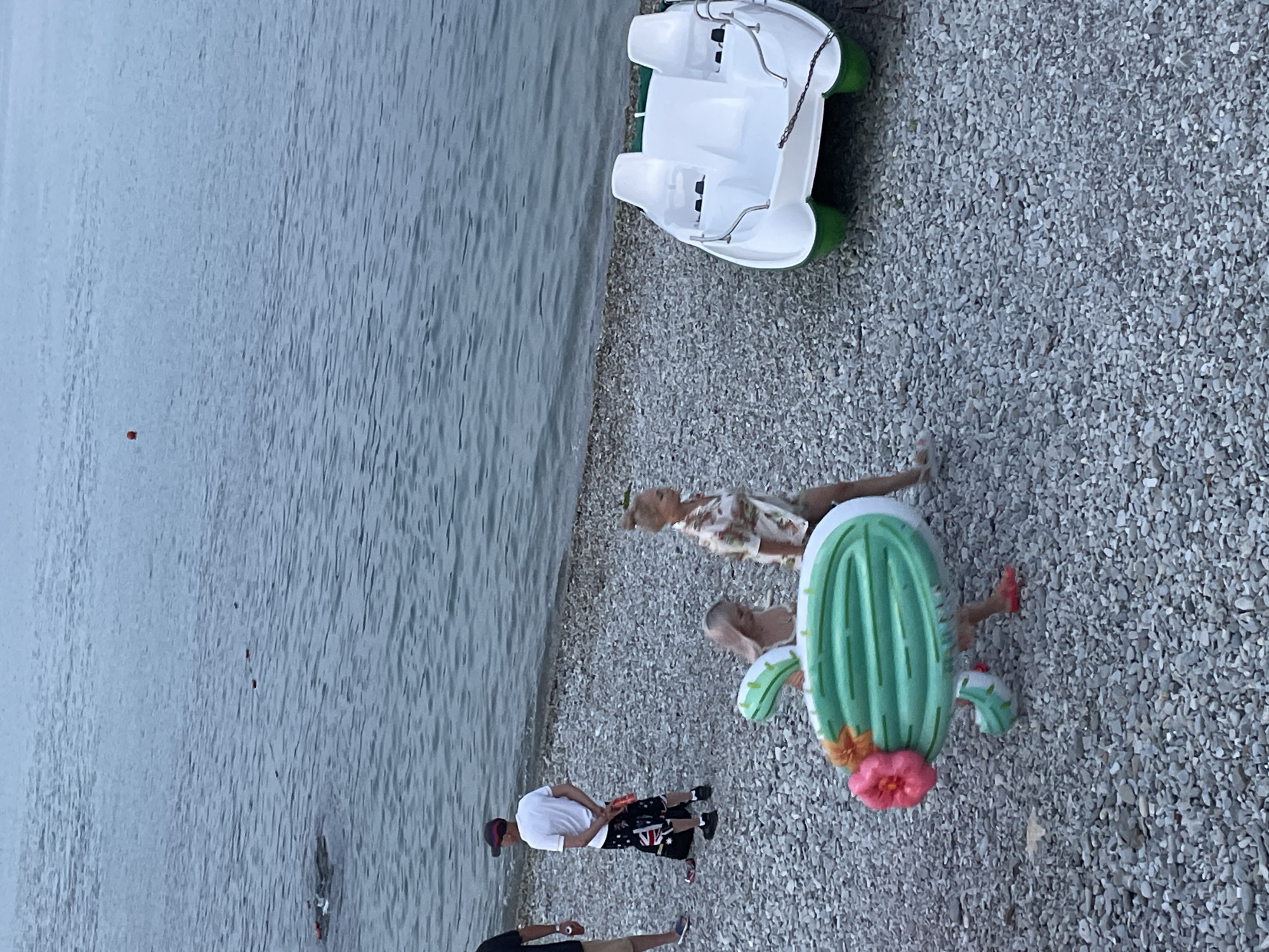 An aerial shot shows a pebble beach with a body of calm water to the left. A white and green pedal boat is pulled up onto the shore on the right. In the center of the frame, two women are walking towards the water. The woman in the foreground is carrying a large, inflatable green cactus with a pink flower. She is wearing a tropical print shirt and shorts. The woman behind her, partially obscured, is wearing a bikini and is being pulled along by the cactus float. To the left, a man in a white t-shirt and patterned shorts stands facing away from the water with his hands behind his back. Another person is partially visible to the far left. There are a few small orange buoys visible in the water. The location is identified as the city of Novorossiysk, Russia.