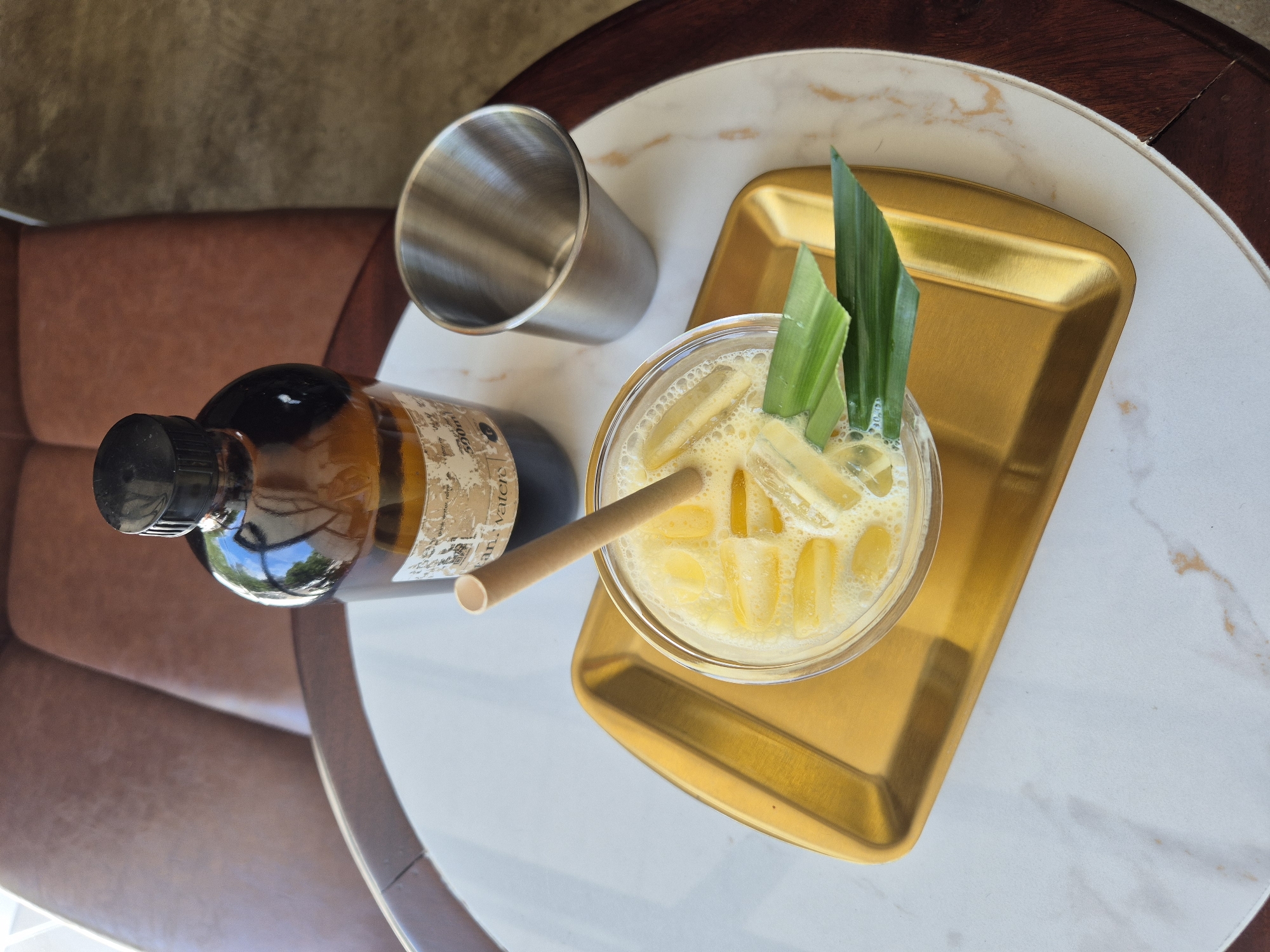 This image depicts a close-up, overhead view of a drink on a gold tray, placed on a marble-patterned tabletop. The drink, in a clear glass, is a light yellow, frothy beverage with ice cubes and a beige straw. Two green pandan leaves are arranged on top of the drink. To the right of the glass is a small, metallic silver cup. Behind the drink, and slightly to the left, is a dark amber glass bottle with a black cap. A label with text is affixed to the bottle. The background features a portion of a brown leather-upholstered chair and a dark wooden table edge. The lighting suggests it is daytime, possibly indoors with natural light contributing to the reflections on the objects. This scene is likely from a café or restaurant in Da Nang, Vietnam, given the presence of pandan leaves, a common ingredient in Vietnamese cuisine and beverages.