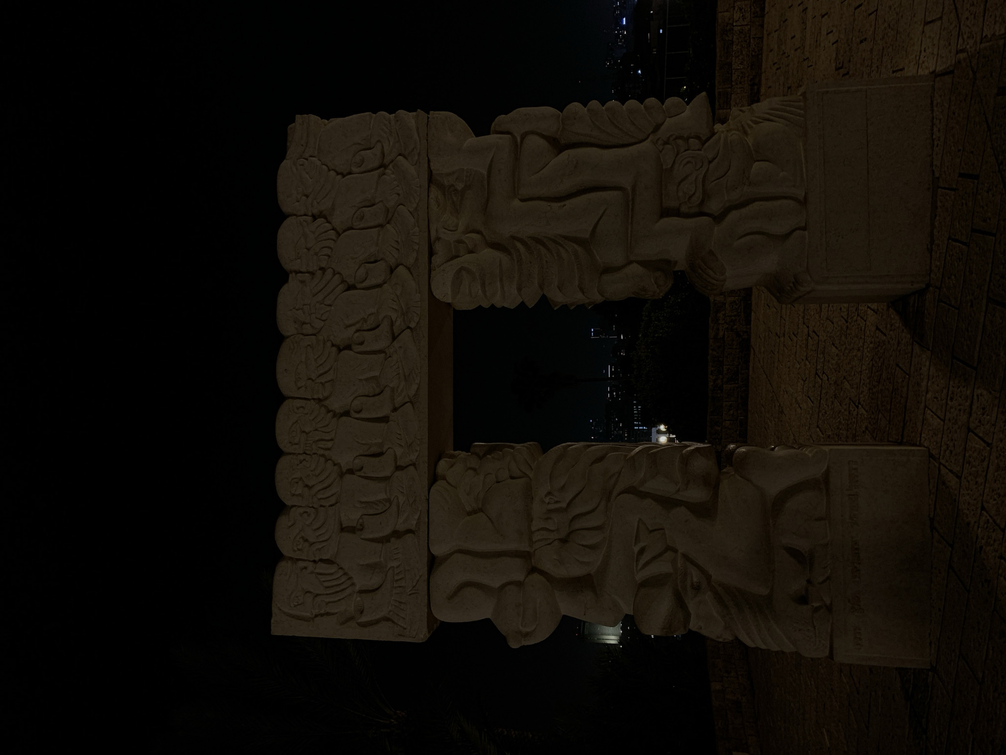 At night, the "Gate of Peace" sculpture, created by Israeli artist Itzhak Danziger in the 1970s, is prominently featured. The sculpture is erected in Tel-Aviv, Israel. The artwork consists of two pillars adorned with bas-reliefs of various human figures. The sculpture is horizontally placed. The foreground is dark, with a wall visible at the right of the shot. The city skyline can be seen through the opening of the sculptures.