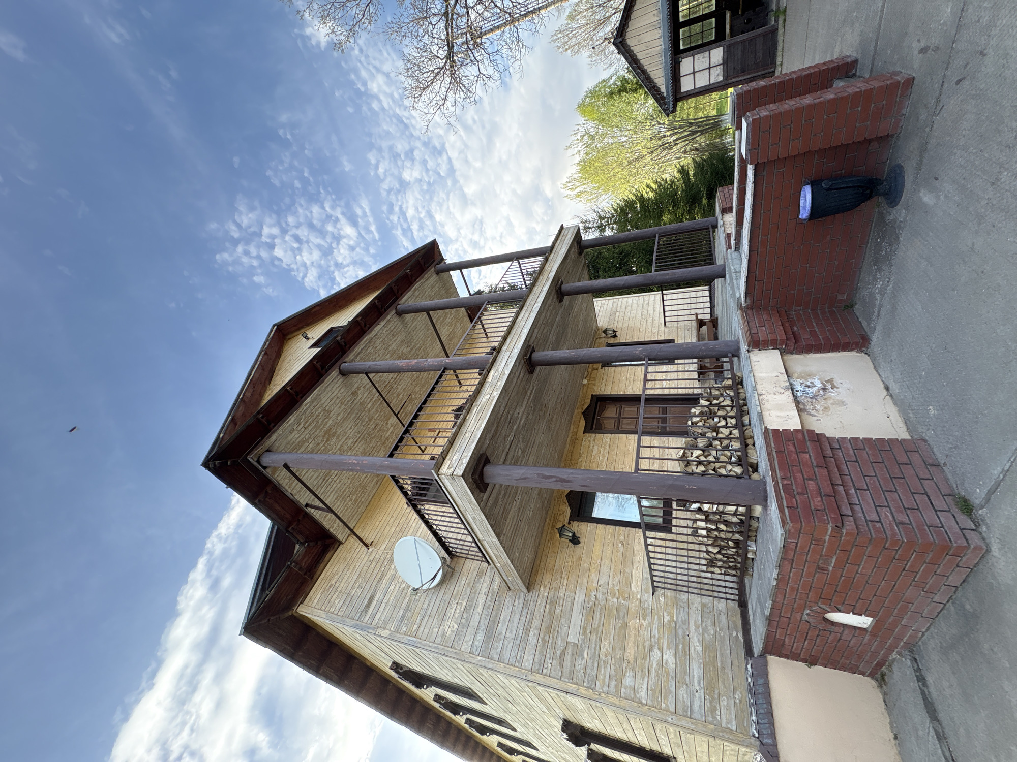 The photograph depicts a three-story house with wooden siding and balconies, located in Zavod Mikhailovskii, Russia. The sky above is blue with scattered clouds. The house features dark brown trim, a satellite dish, and cylindrical posts supporting the balconies. A brick structure with a black urn sits to the side. Firewood is stacked on the ground floor balcony.