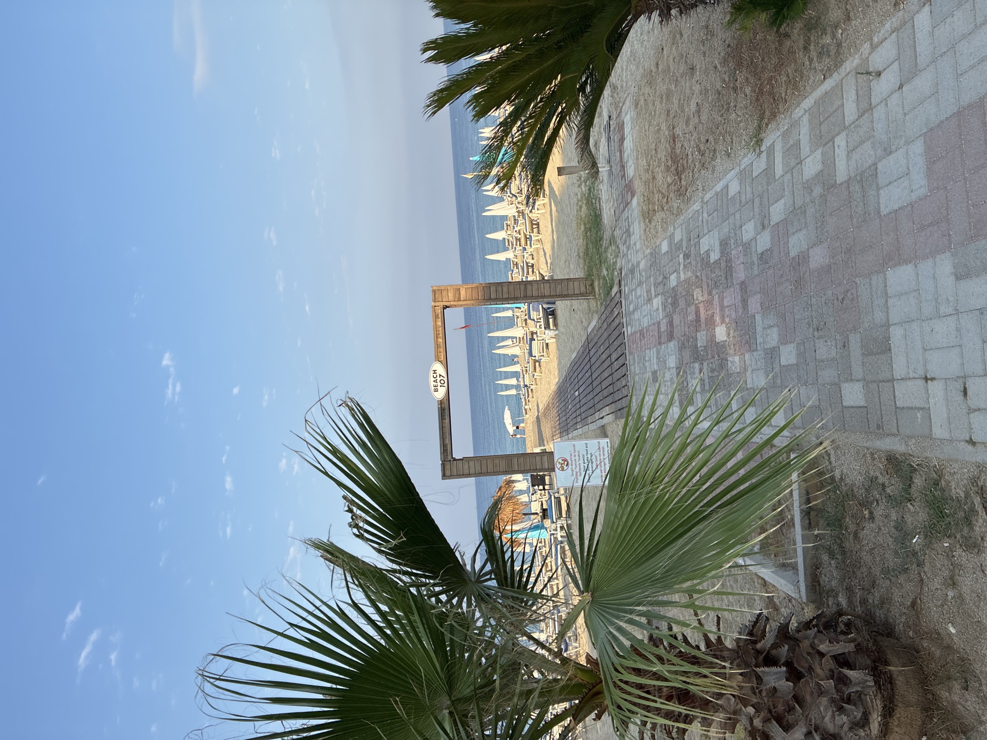 The image shows an entrance to Beach 107 in Alanya, Turkey. The entrance is marked by a wooden archway, above which a sign reads "BEACH 107". A wooden pathway leads through the archway to a sandy beach lined with rows of white lounge chairs and umbrellas. The sea is visible in the background under a clear blue sky with a few scattered clouds. Two palm trees frame the entrance, one on the left and one on the right. A sign is posted near the entrance with text in multiple languages, likely a set of beach rules or regulations. The overall scene depicts a well-maintained beach resort area.