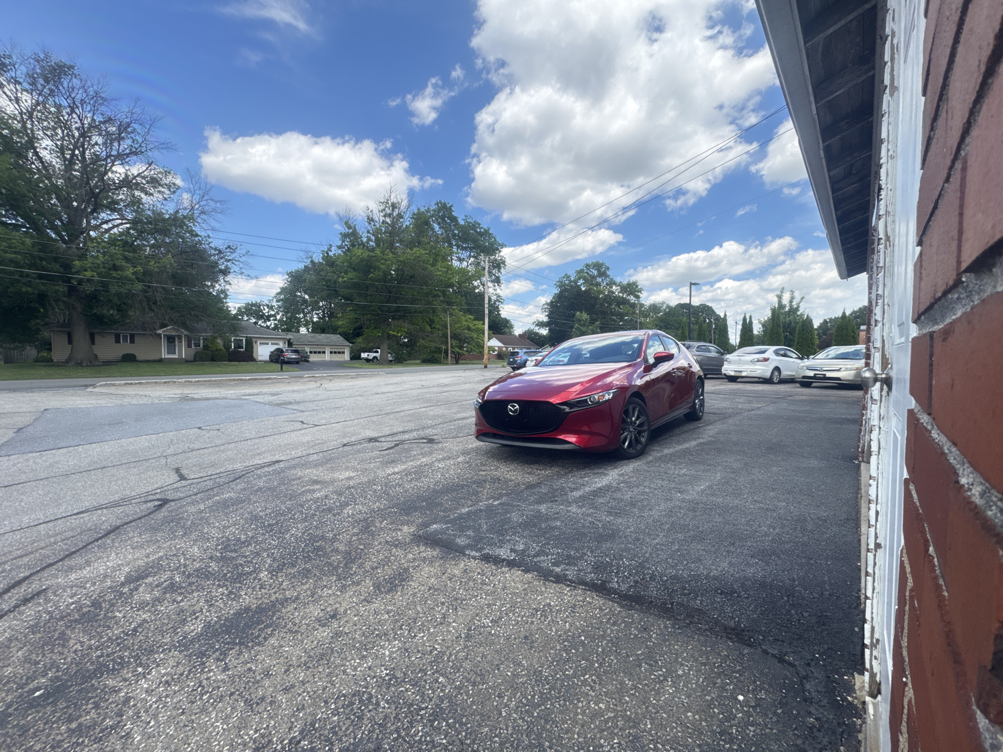 A red Mazda 3 is parked in an asphalt lot in front of a brick building with a white door. The car is facing right. In the background, there are several other vehicles parked, including a white Honda sedan and a silver Honda sedan. Behind the cars is a two-story building with a beige facade and a dark roof. A large tree with green foliage is to the left of the building. The sky is blue with scattered white clouds. The image is taken from a low angle, with the front of the car prominently in the foreground. The scene appears to be a car dealership or parking lot.