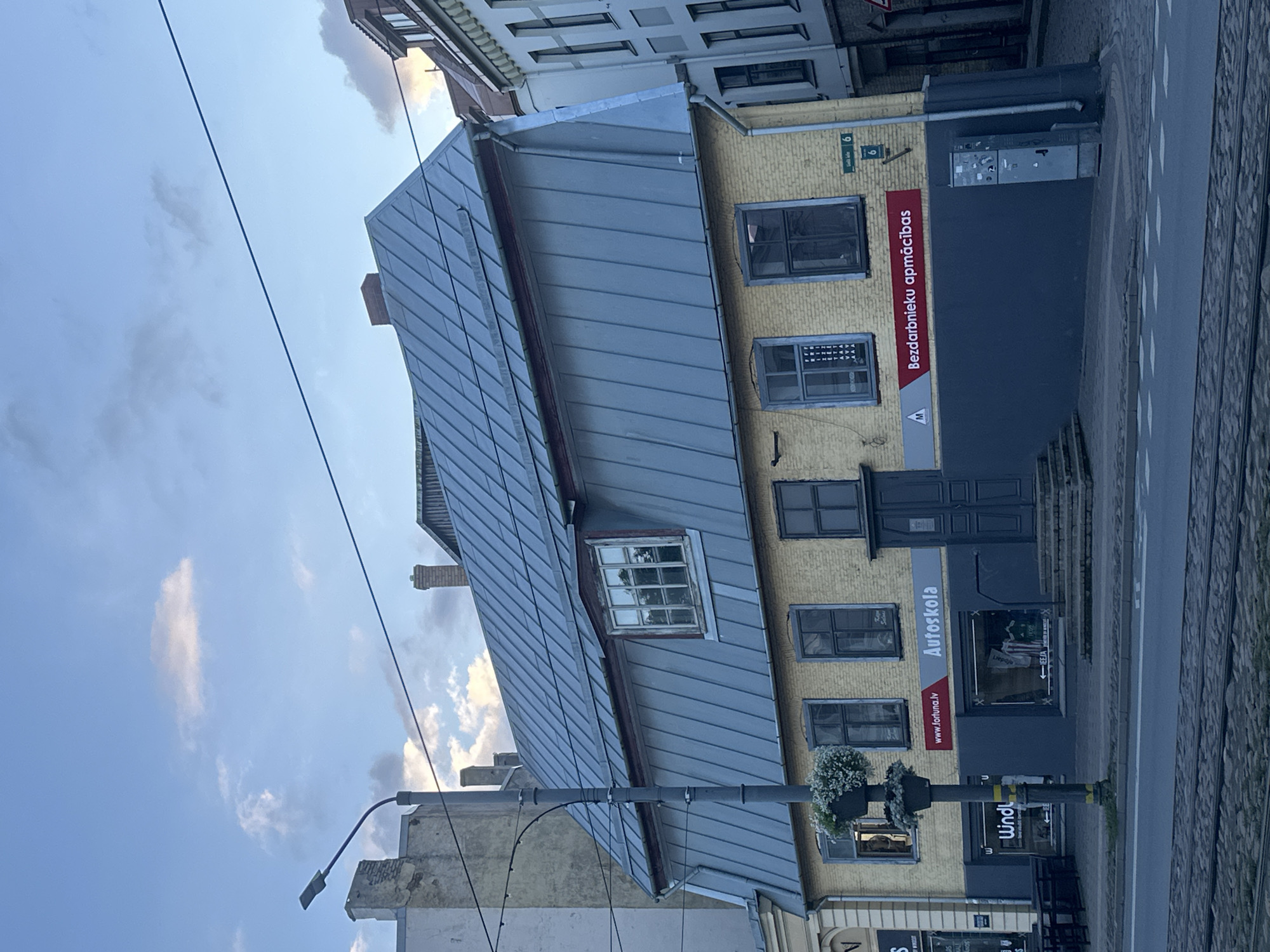 This is a daytime, eye-level, slightly low-angle exterior photograph of a yellow brick building with a grey, mansard-style metal roof. The building is located on a street with tram tracks in the foreground. The building has multiple windows on its upper level and appears to be a commercial establishment on its lower level. Signs on the building indicate it houses an "Autoskola" (driving school) and "Bezdarbnieku apmācības" (unemployment training). There is a street lamp with hanging flower baskets in front of the building. The sky is partly cloudy with a mixture of blue and grey. The architecture suggests an older European city. Based on the signs and architectural style, the location is likely in Liepaja, Latvia. There are no people visible in the image.