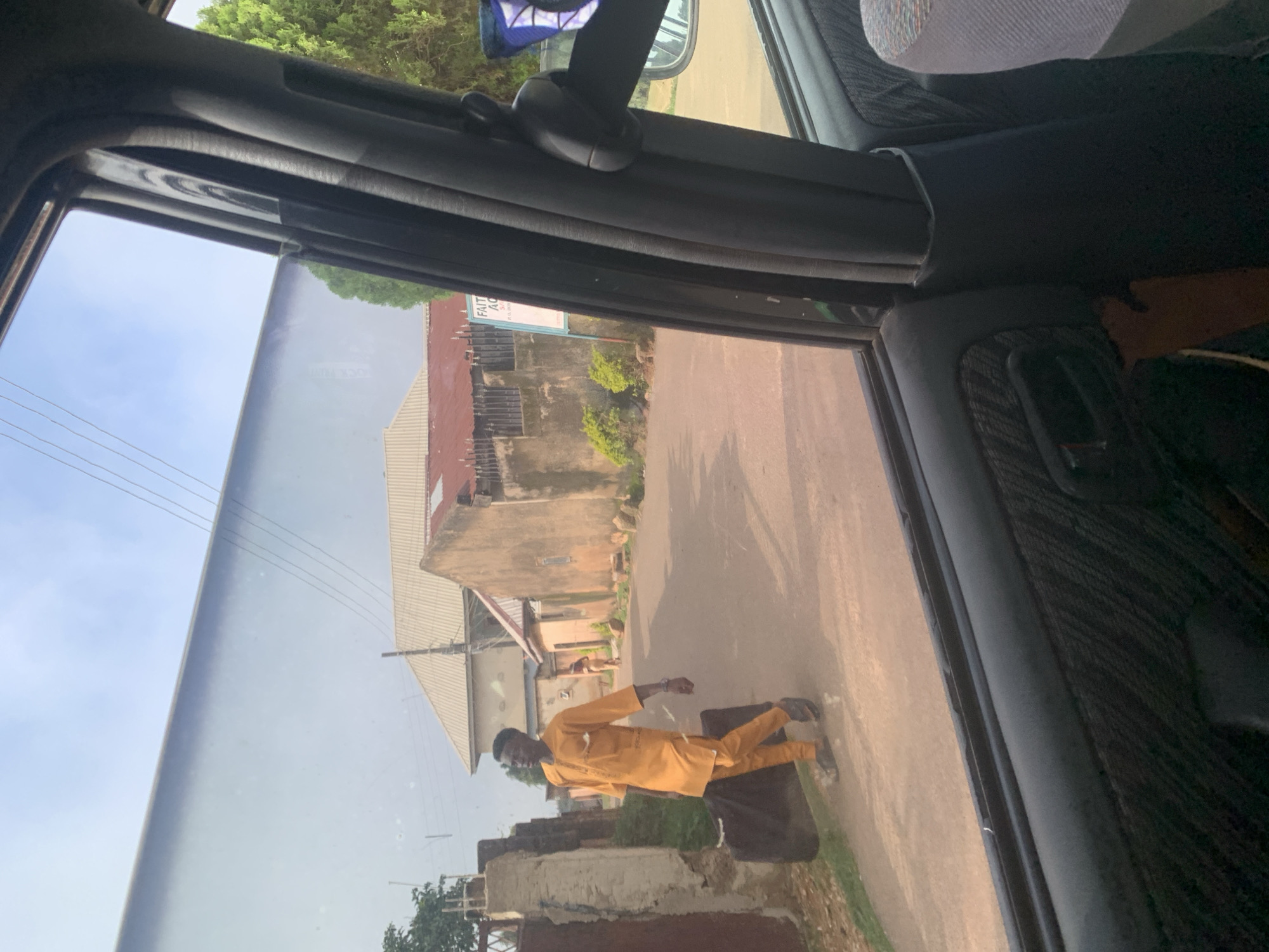A man in an orange traditional outfit walks on a street in an urban area, pulling a dark suitcase. He is seen from the passenger side of a vehicle, looking towards the left of the frame. The street is paved, and there are buildings on either side. Some buildings appear to be residential, with corrugated metal roofs and concrete walls. There is vegetation visible, including trees and bushes. The sky is partly cloudy. The interior of the vehicle is partially visible, including the car door panel, a seat, and part of the dashboard. The scene appears to be during daylight.