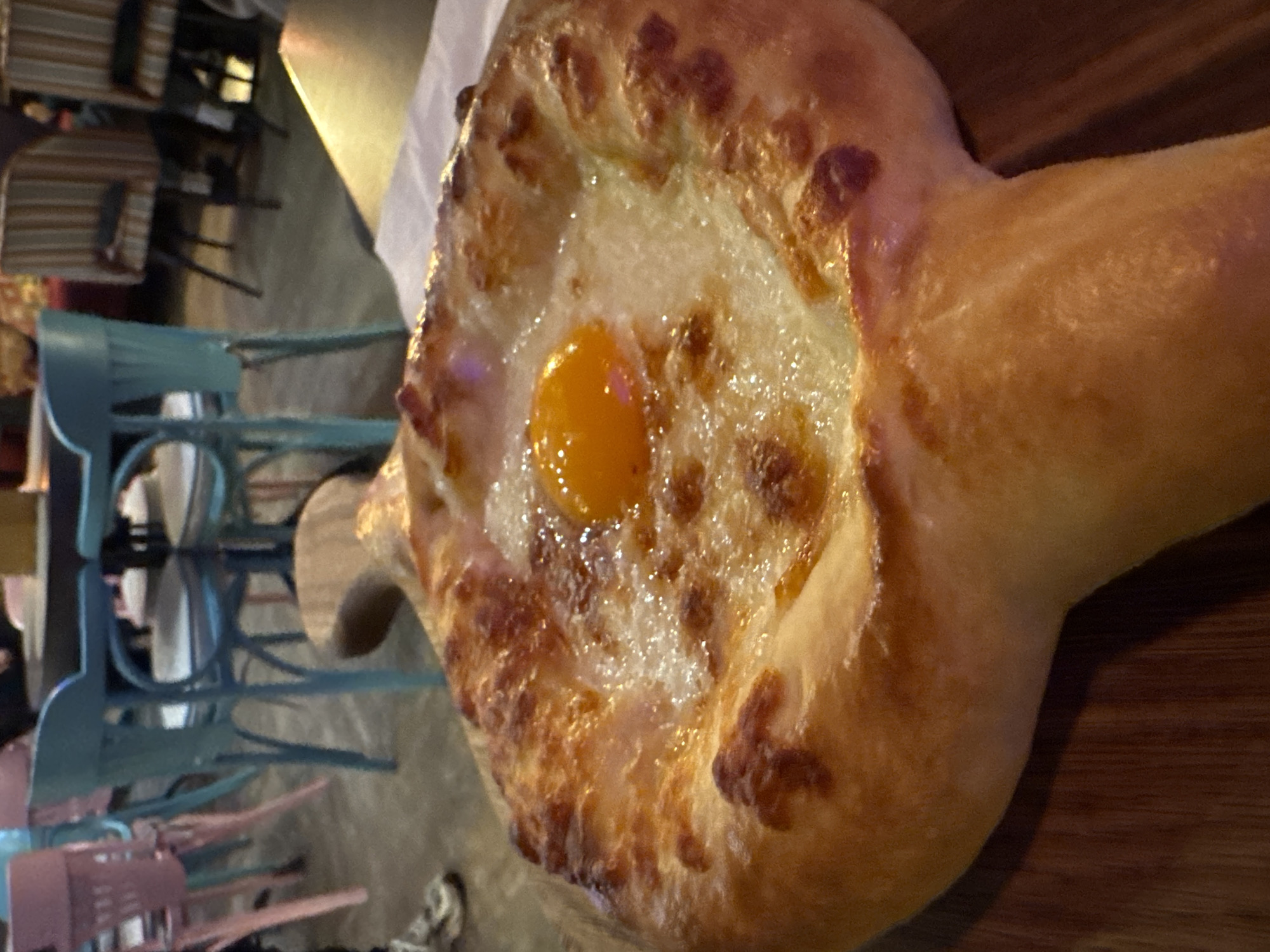 A close-up photograph shows a khachapuri, a traditional Georgian dish, sitting on a wooden board. The khachapuri is boat-shaped, with a baked crust and a filling of cheese and an egg yolk in the center. The crust is golden brown and slightly charred in places. The cheese appears melted and bubbly, and the egg yolk is a vibrant orange. In the background, partially out of focus, there are tables and chairs, suggesting the scene is inside a restaurant. The chairs are a mix of teal and pink. The location is Saint Petersburg, Russia.