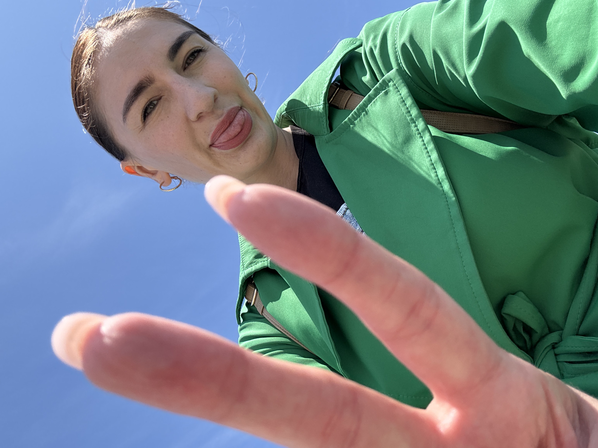 A woman in a green jacket is shown from a low angle, sticking her tongue out and making a peace sign. The background is a clear blue sky. There is no information provided in the image about the city of Sochi, Russia.