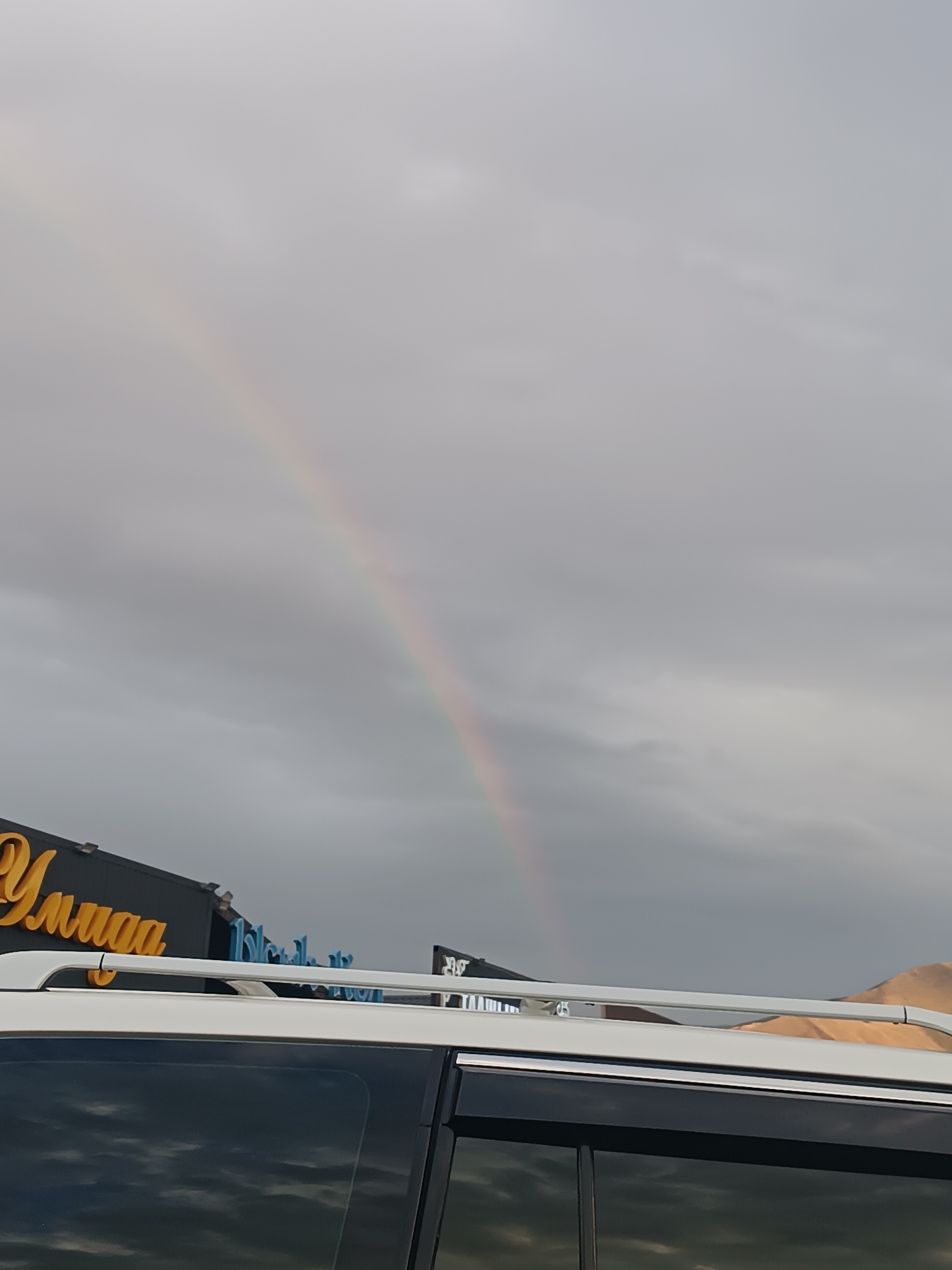 An overcast sky with a visible rainbow arching across it is captured from the perspective of a vehicle's roof rack. In the lower left portion of the image, a dark building with a yellow sign that reads "Улица" (Ulytsa) is visible. To the right of this sign, a blue neon sign is partially obscured. The foreground shows the roof and upper portion of a white vehicle, with its tinted windows reflecting the sky. The environment appears to be outdoors in the city of Sasyk-Bulak, Kyrgyzstan.