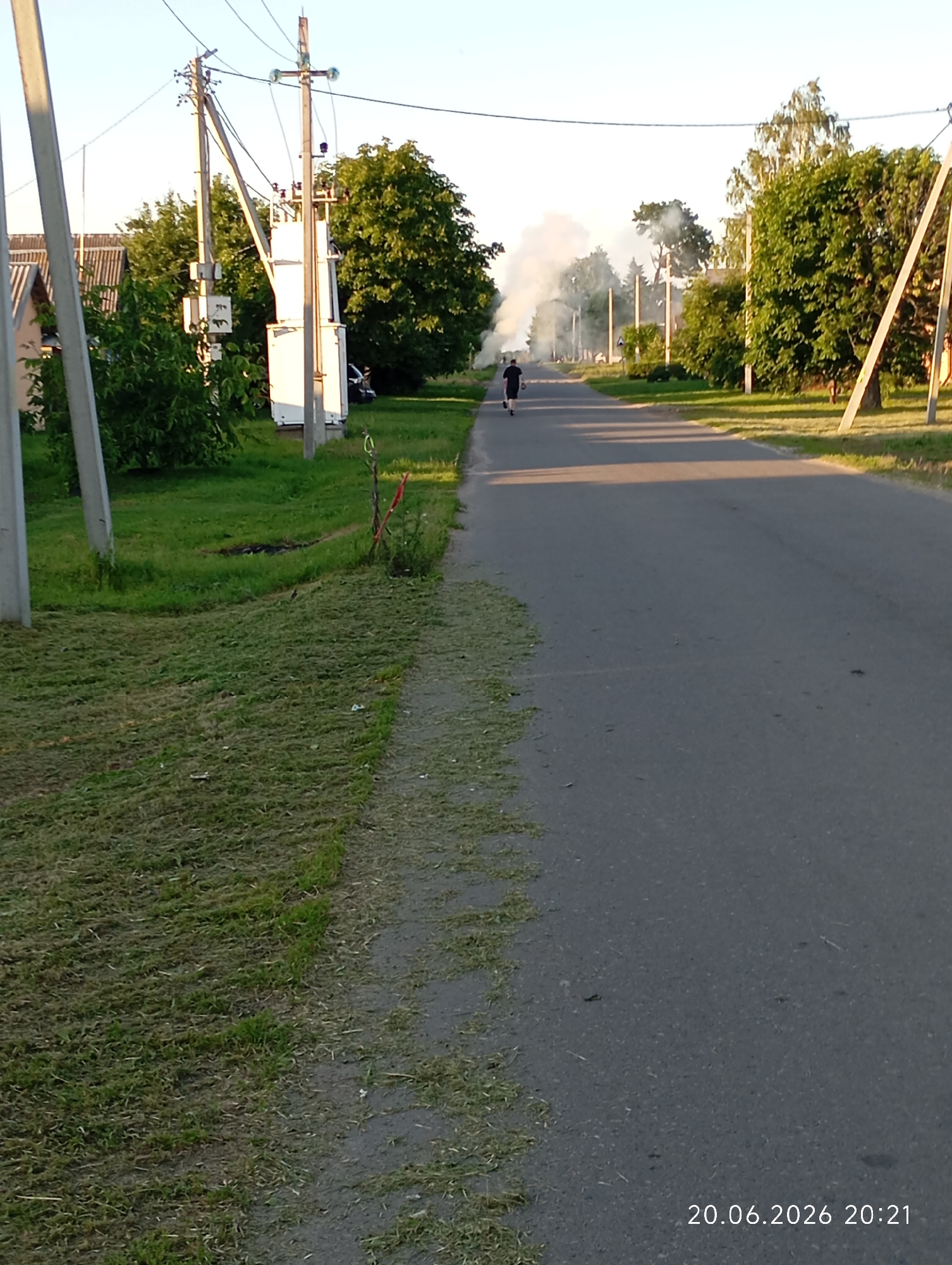 The image shows a rural road in Урыцкі сельскі Савет, Belarus, with a person walking away from the camera. Dense white smoke is rising from an area further down the road, suggesting a fire. To the left of the road, there is a grassy area with freshly cut grass. Utility poles and trees line the road, and houses are visible in the background. The time stamp on the image indicates it was taken on June 20, 2026, at 20:21.
