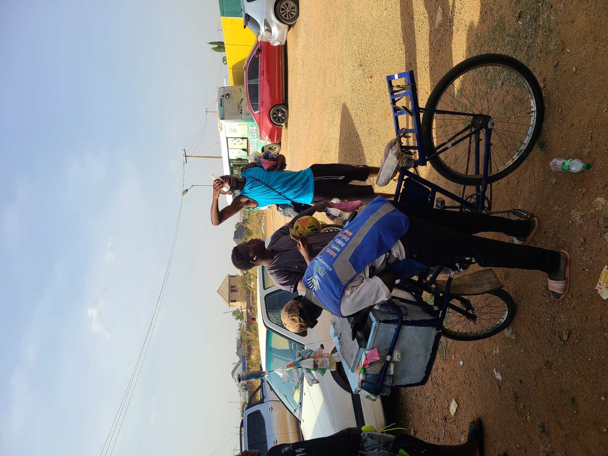 Three men are gathered around a bicycle that is heavily loaded with goods. The men are standing on a dirt surface, with parked cars and buildings visible in the background, suggesting the edge of a commercial or residential area. The bicycle is blue and is positioned next to a white car. The location is in New Stadium, Nigeria. One man is wearing a blue shirt and holding a water bottle near his head. Another man is holding a yellow and green object. The third man is positioned near the white car and the bicycle.