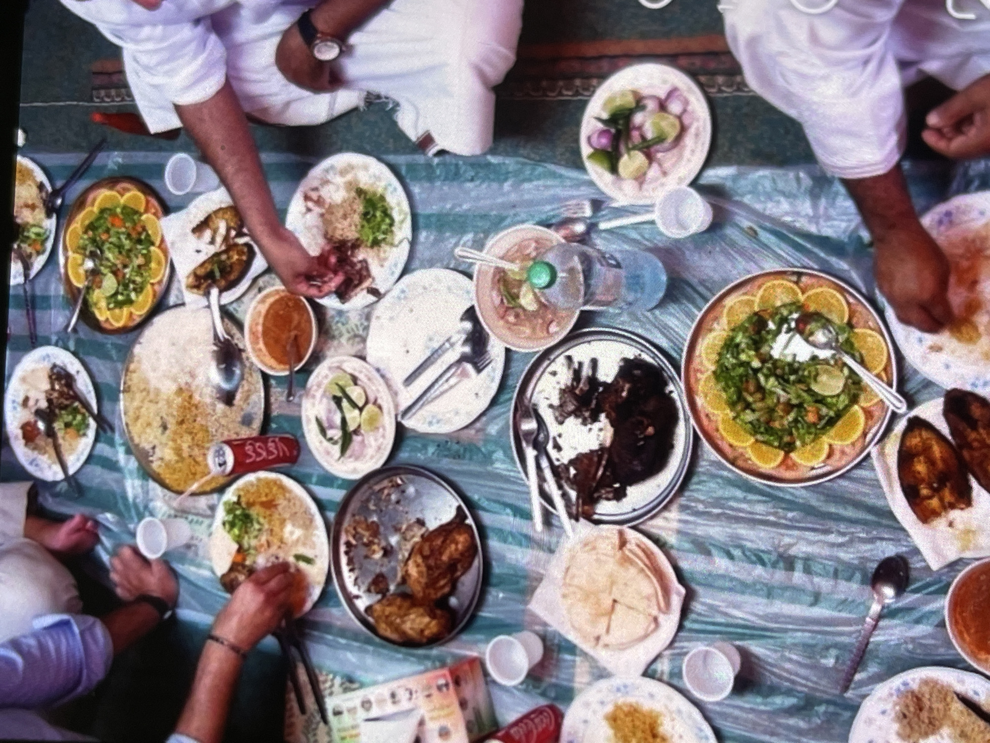 The image shows an overhead view of a group of people seated on the floor around a spread of food. The food is laid out on a plastic tablecloth, which is spread on the floor. There are several plates of rice, meat, salad, and bread. A can of Coca-Cola is also visible. People's hands are visible reaching for the food. The scene appears to be a casual meal.