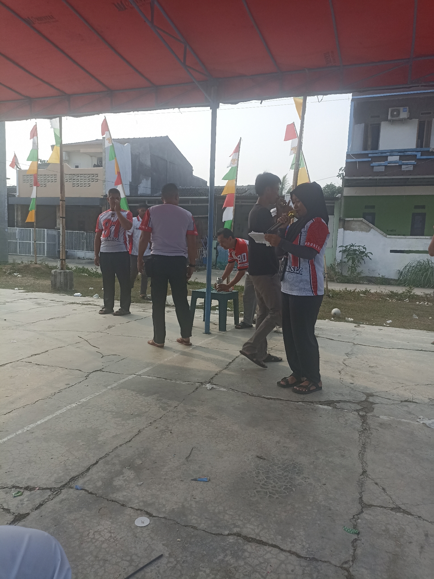 The image depicts an outdoor event in the city of Cibarusah, Indonesia, likely a community gathering or competition held on a cracked concrete court. Several people are present, some actively participating and others observing. In the foreground, a woman wearing a hijab and a patterned shirt is holding a microphone and a piece of paper, seemingly addressing the group. To her left, a man in a black shirt stands beside her, also holding a paper, possibly another speaker or an assistant. Behind them, another man in a red and white jersey with the number "87" is visible, looking towards the speakers. Further back, a person in a light purple shirt is also present. The scene is partially covered by a red awning. Flags, including red and white Indonesian flags, are planted in the background. Buildings and trees are also visible in the distance, suggesting a suburban or semi-rural setting. The ground is littered with some debris, including white papers and plastic wrappers. The overall impression is of a casual public event taking place during daylight hours.