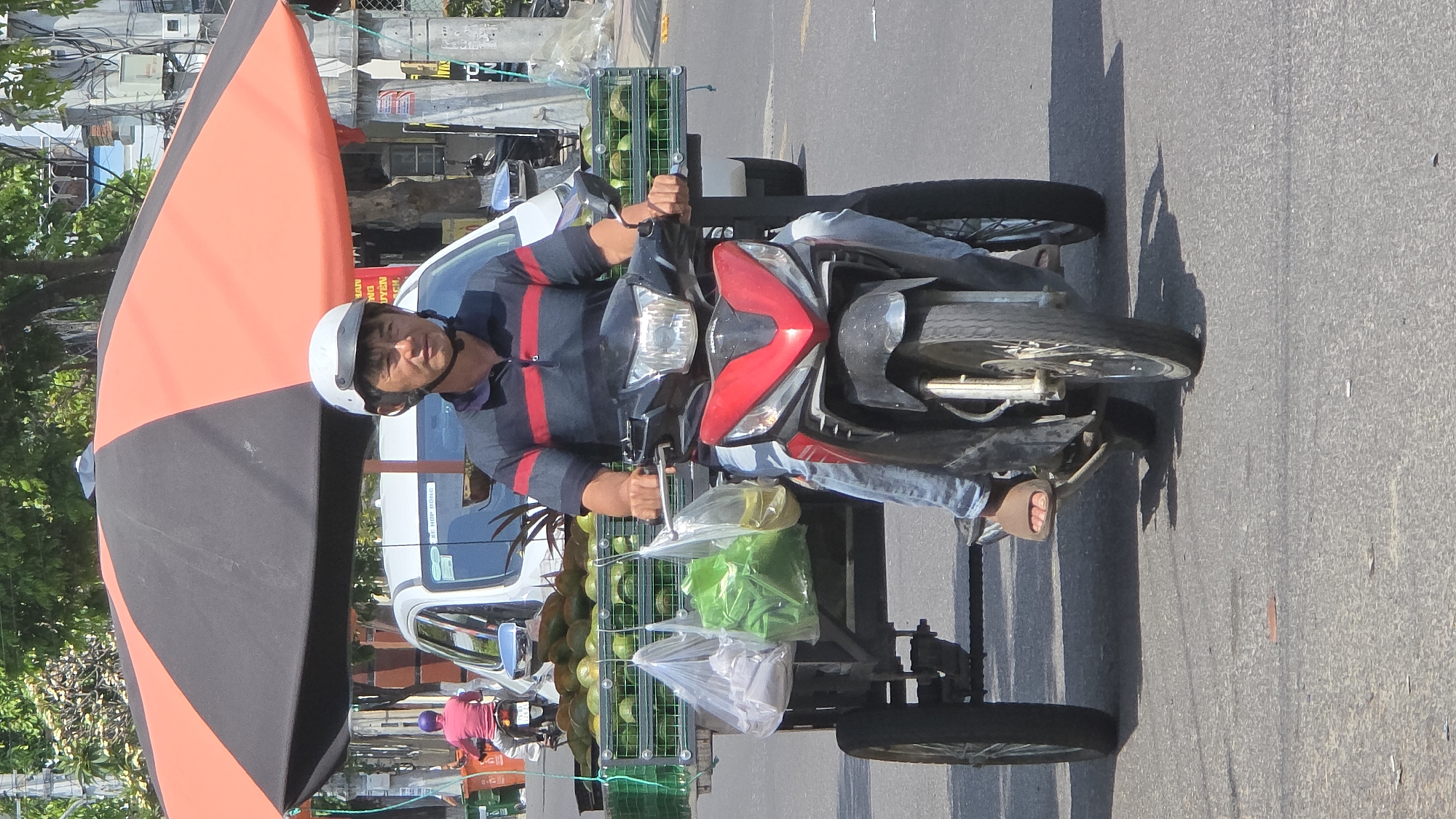 A man is riding a motorcycle with a sidecar attached to it. The sidecar is carrying a load of melons, and he also has a large umbrella attached to the motorcycle. He is wearing a helmet and casual clothing. The scene appears to be on a street in Nha Trang, Vietnam.