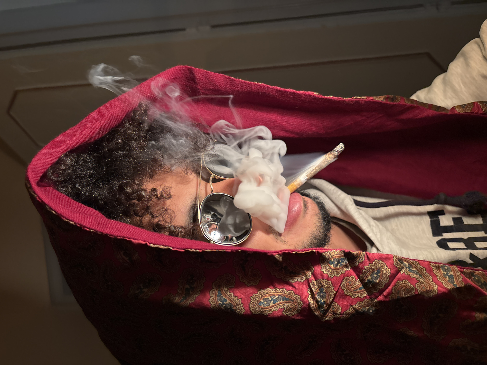 A person is lying down, wearing round sunglasses, smoking, and appears to be enveloped in a red patterned fabric. The location cannot be determined from the image.