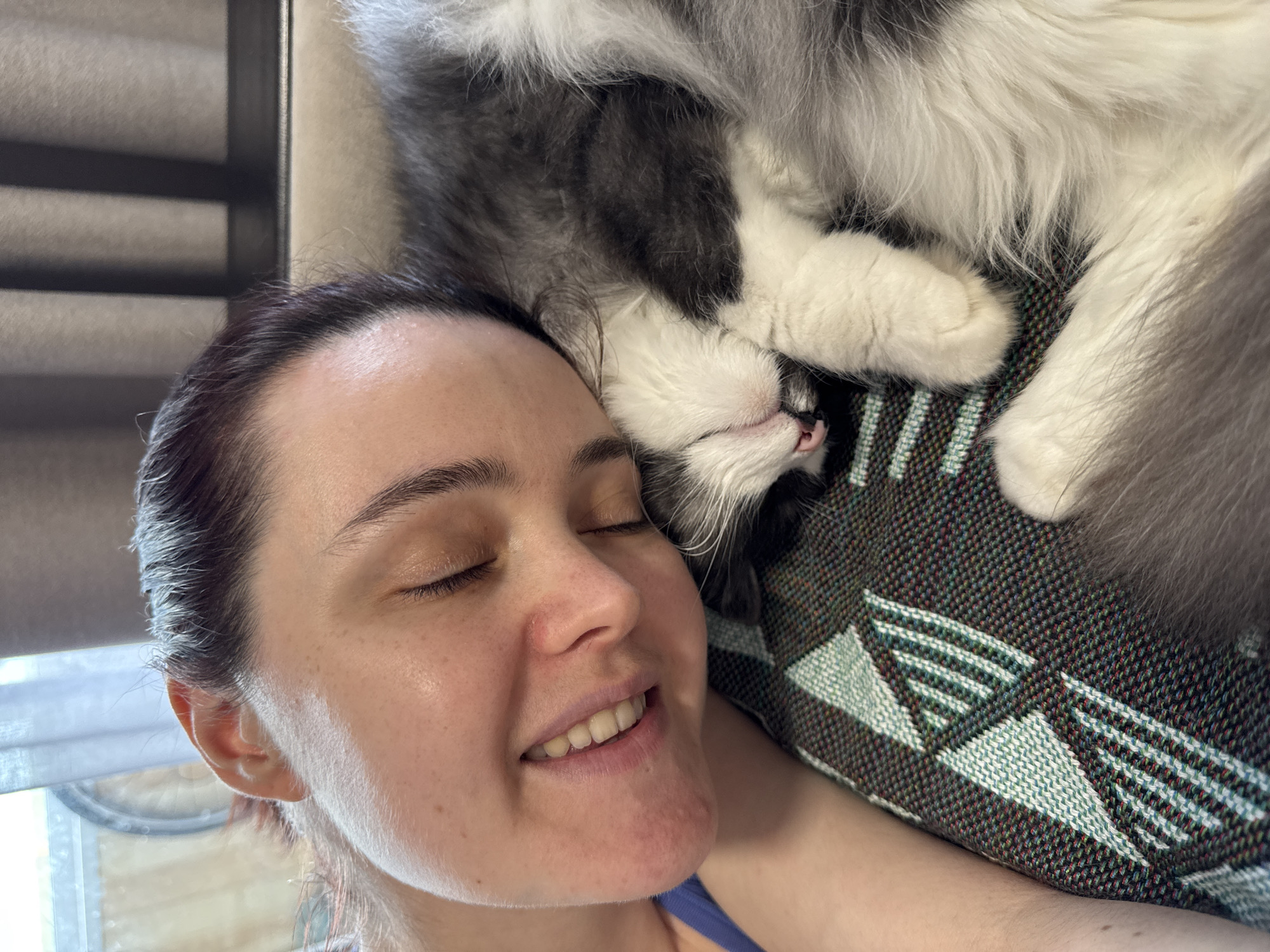 A woman with dark hair pulled back from her face is lying down with her eyes closed, smiling slightly. A large gray and white cat is lying on top of her head and upper body. The woman is wearing a blue tank top. She is lying on a patterned gray, green, and white woven blanket. A window with horizontal blinds is in the background. There is no indication that this image was taken in Izhevsk, Russia.