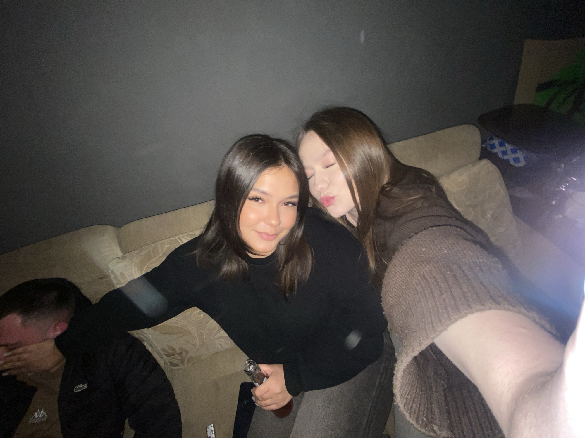The image depicts three young individuals gathered in a dimly lit indoor setting, likely a casual social event or party in Chaikovsky, Russia, during the evening or nighttime. In the foreground, a young woman with dark, shoulder-length hair smiles subtly, looking towards the viewer. She wears a black long-sleeved top and dark pants, with a nose piercing visible. She holds a brown glass bottle. Leaning into her from the right, another young woman with long, light brown hair poses playfully, with closed eyes and a "kissy face," seemingly taking the photo as a selfie. She is dressed in a brown, possibly knitted, long-sleeved top.
To the far left, partially obscured, a man with short hair covers his face with his hand. He wears a dark puffer jacket with a white Lacoste crocodile logo, over a lighter-colored shirt with indistinct white text. All three are seated on or leaning against a light-colored, patterned sofa. The background features a plain dark gray wall. In the upper right, a dark bag with a blue and white patterned item, and a dark chair draped with a bright green object, are visible. The scene is brightly illuminated by a camera flash, highlighting the subjects against the dark environment. The overall mood appears casual and friendly.