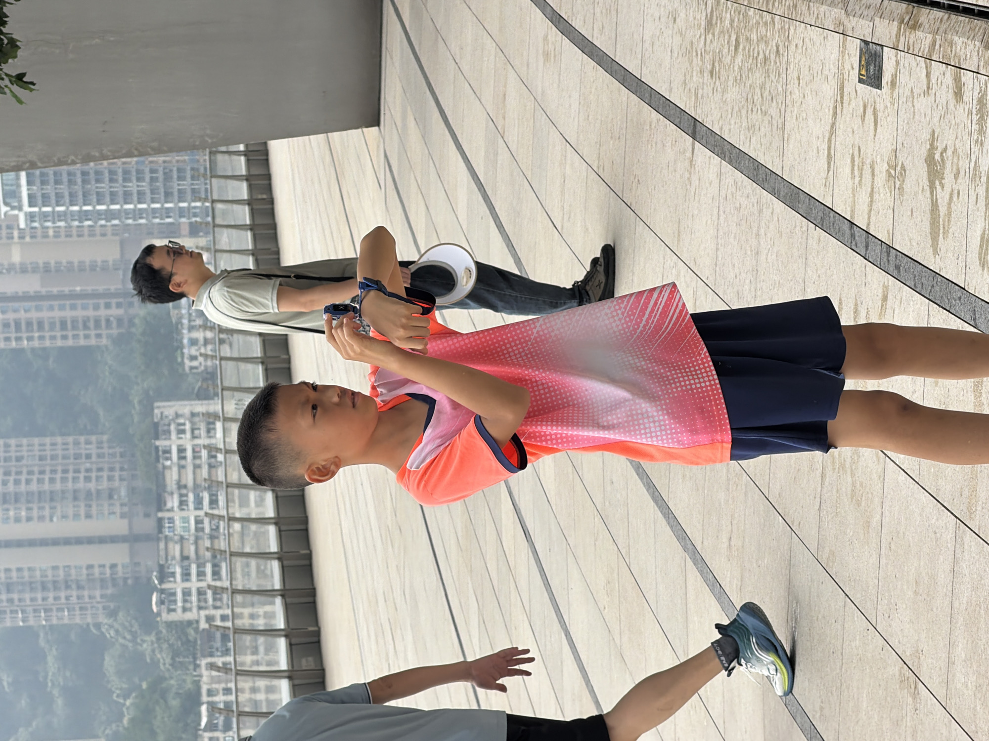 The image captures a scene in Yuzhong District, China, featuring three individuals on an elevated pedestrian walkway with buildings visible in the background. In the foreground, a young boy with a close-cropped haircut is wearing an orange and pink patterned t-shirt and dark blue shorts. He is holding a blue electronic device, possibly a camera or a watch, in his hands and looking intently at it. To the left of the boy, another person, partially visible from the waist down, is wearing a light blue t-shirt and black shorts, with one foot in a blue and white athletic shoe. They are walking away from the viewer, with their left hand extended. Further in the background, a man wearing glasses, a light green polo shirt, and dark jeans is seen standing. He has a dark bag slung over his shoulder and is holding a white disc-shaped object. The walkway is paved with light-colored tiles and divided by dark gray strips. The environment appears to be an urban setting with buildings and a hazy sky.