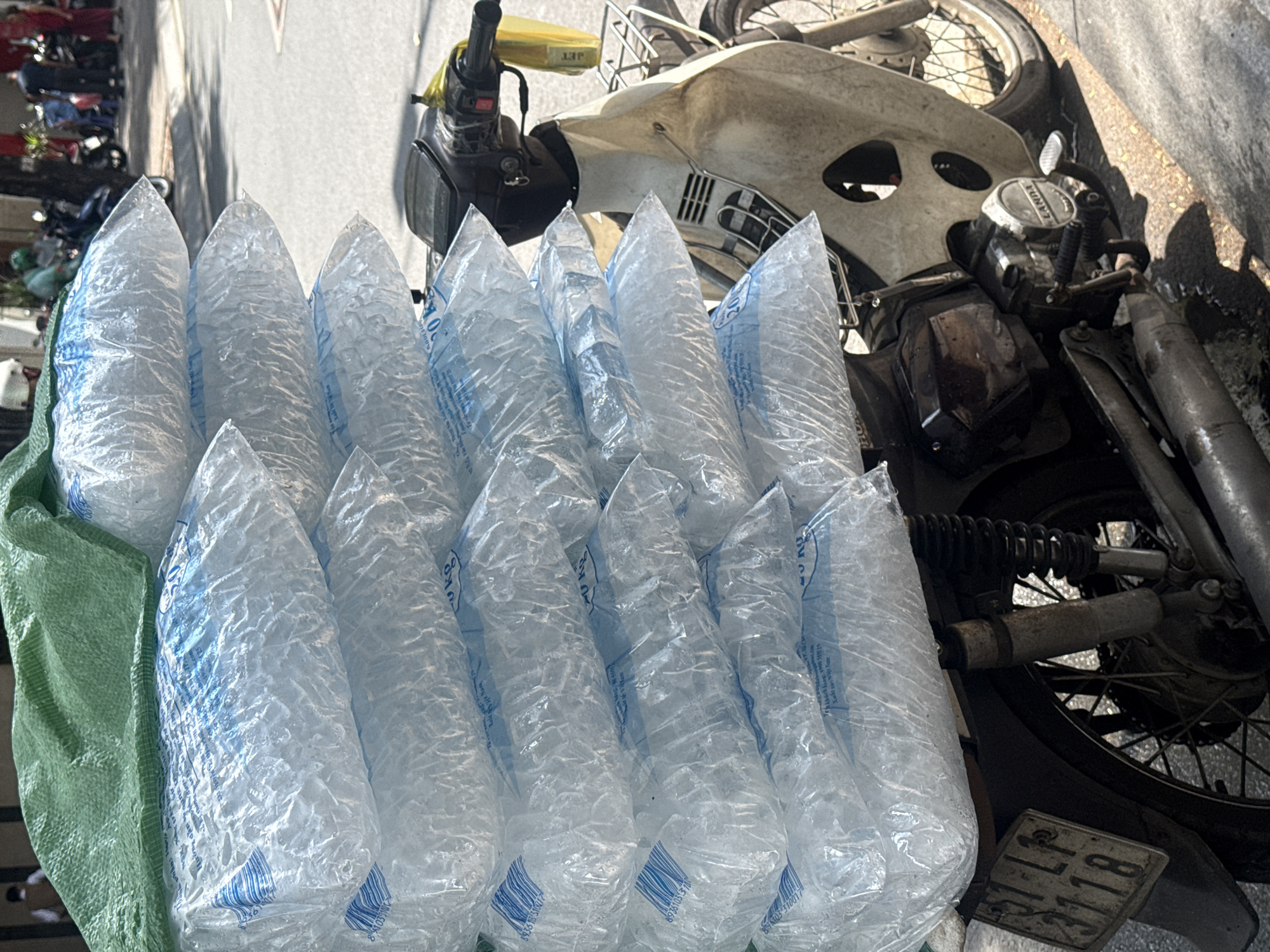 A close-up, eye-level shot shows a stack of large, transparent plastic bags filled with crushed ice, piled high on the back of a beige, weathered motorcycle. The bags are printed with blue lettering and barcodes. A green tarp is draped over the top of the ice bags. The motorcycle's handlebars, front and rear fenders, and a worn license plate are visible. The license plate reads "51-LP 3118". The motorcycle is parked on a paved street with a sandy or gravelly edge. In the blurred background, people and other motorcycles are visible, suggesting an urban environment. This scene appears to be in Thủ Đức, Vietnam, a city known for its busy streets and commerce.