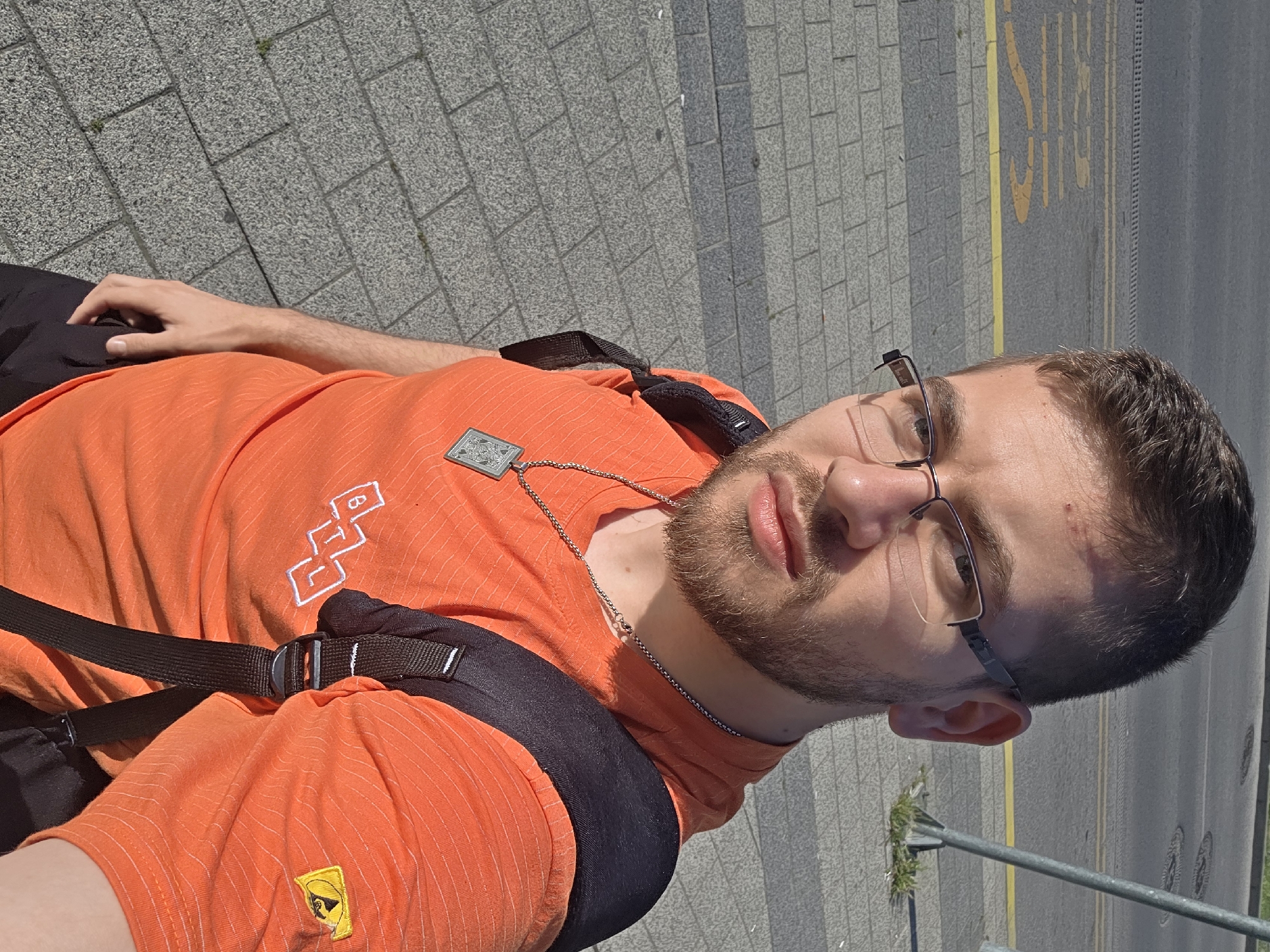 A medium shot depicts a man wearing an orange t-shirt and glasses. He has a beard and is wearing a backpack. The t-shirt features a white geometric design and a yellow patch. He is wearing a silver necklace with a pendant. The man is standing on a paved surface with a yellow painted line near a road. This is likely a sidewalk or pedestrian area in Plovdiv, Bulgaria.