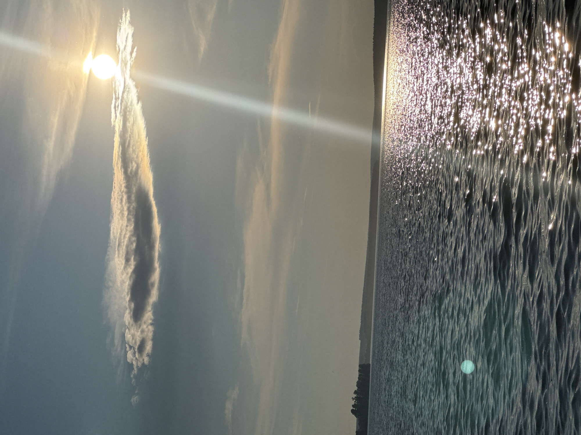 The image depicts a serene natural scene, likely a body of water in Votkinsk, Russia, during daylight. The sky is a pale blue with scattered clouds. A prominent, elongated cloud is positioned in the upper portion of the frame, with the sun partially obscured behind it. The sun casts a bright beam of light downwards, reflecting intensely off the surface of the water. The water itself appears calm, with gentle ripples, and is a deep blue-green color. A distant shoreline is visible, characterized by a dark line of trees against a hazy horizon. There are no people or man-made objects immediately apparent in the scene. The overall atmosphere is peaceful and natural.