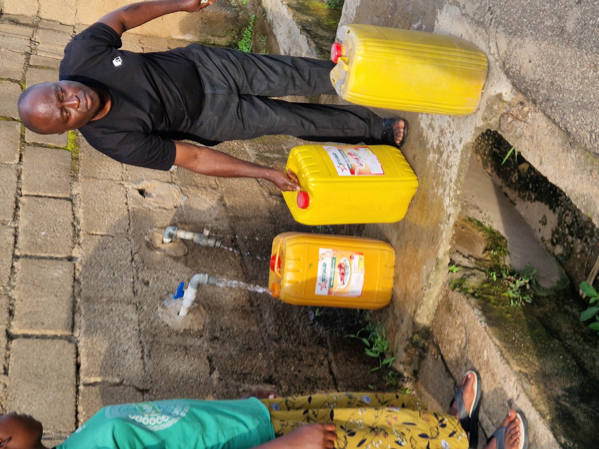 In Kabong, Nigeria, a man in a black t-shirt and dark pants is seen at a water source. He is holding a large yellow plastic container with a red lid, which appears to be filling with water from a tap. To his left, another similar yellow container is positioned under a running tap. A younger person, partially visible on the left side of the frame, wears a green t-shirt and a patterned yellow skirt, observing the water collection process. The background consists of a rough brick wall. Water is flowing from the tap into one of the containers, and there is a drainage channel in the foreground where water is visibly running. Three yellow containers, two of which are actively involved in water collection, are prominent in the scene.