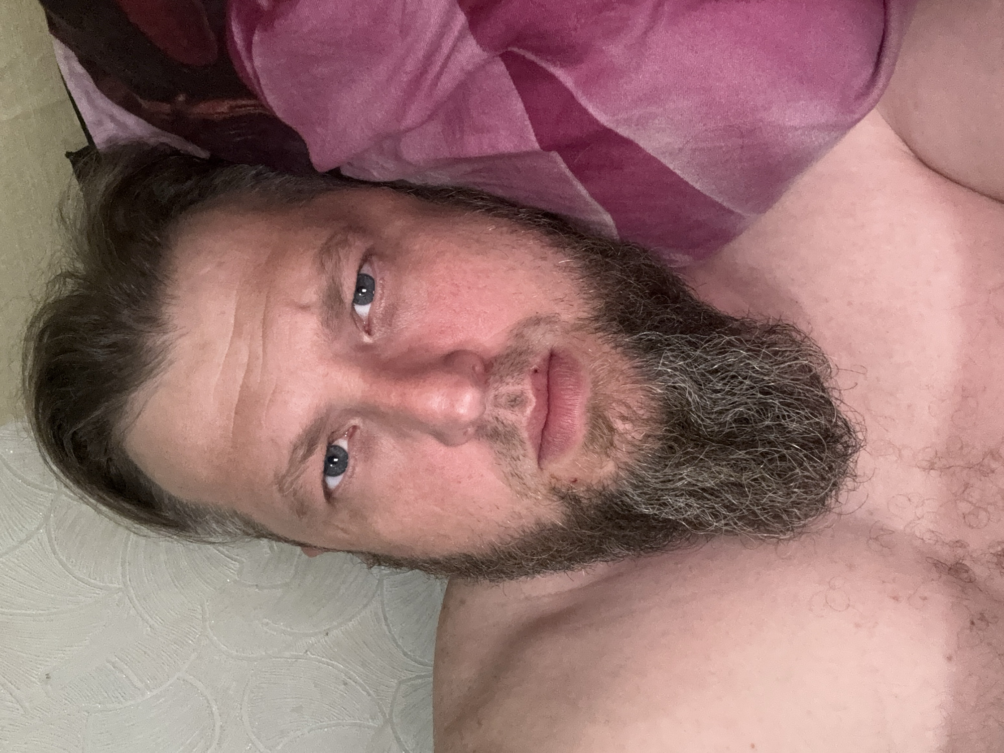A man with a beard and blue eyes is lying in bed, looking at the camera. He is wearing a pink blanket. The room is dimly lit and the wall behind him has a textured pattern. The image appears to be a selfie taken indoors.