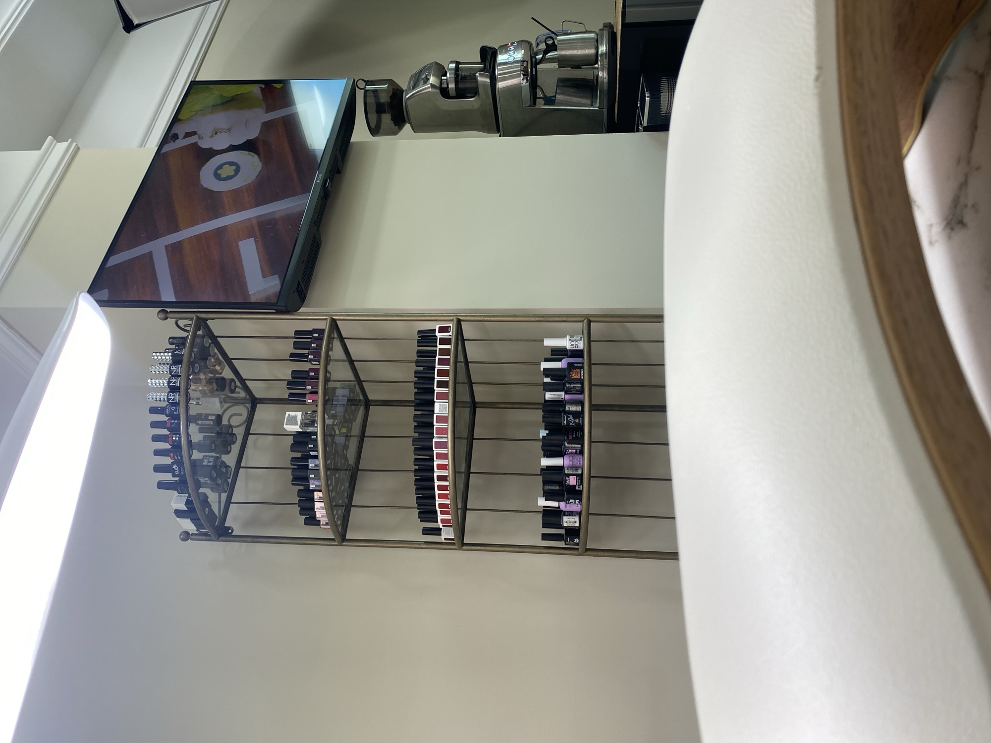 This appears to be the interior of a nail salon. On the wall, there is a TV screen playing a video. Above that, there is a coffee machine. Below the TV, there is a wall-mounted shelving unit displaying a variety of nail polish bottles arranged by color. A white chair is visible in the foreground. The location is unknown and not discernible from the image.
