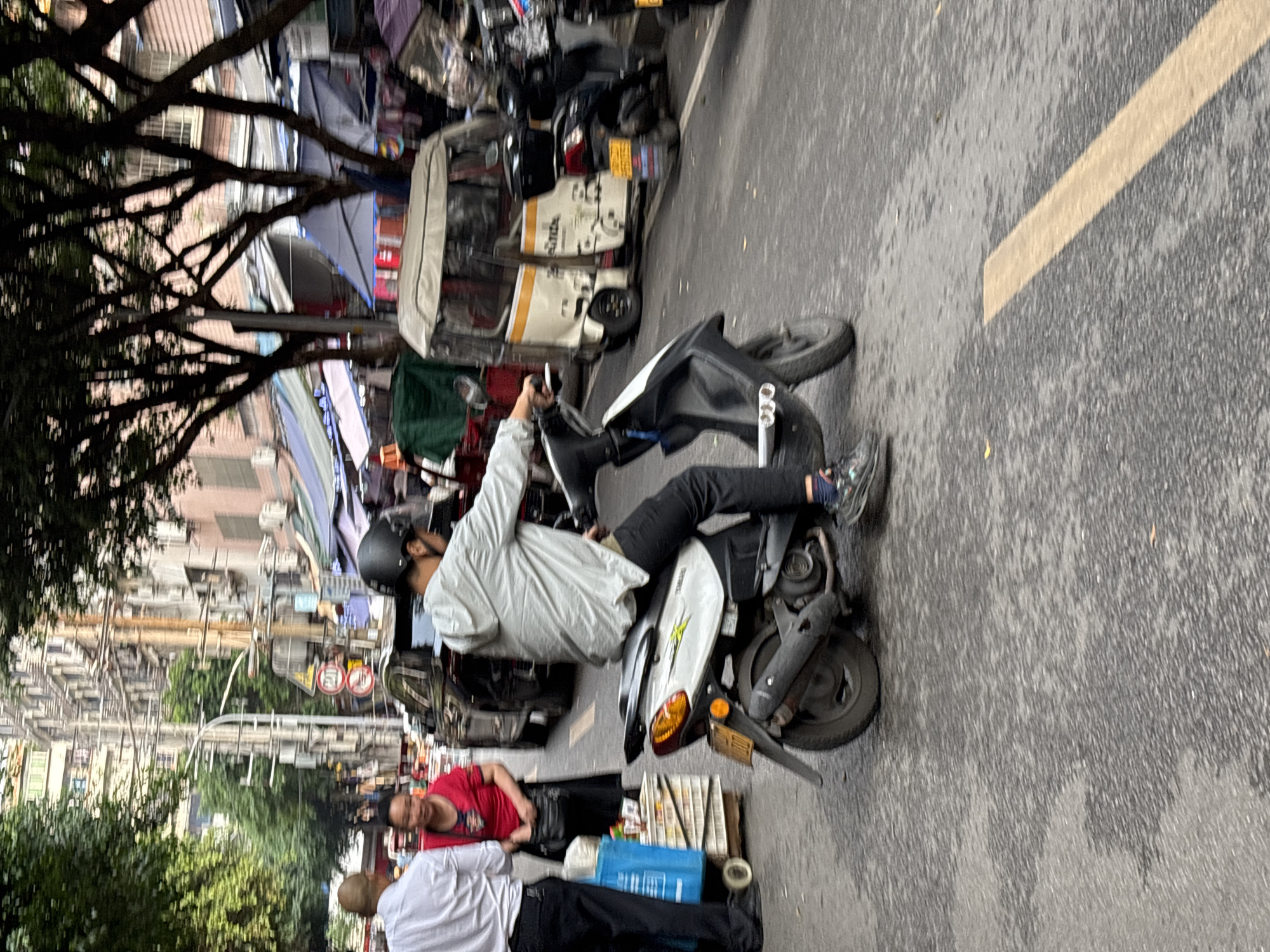 The image depicts a street scene in Jiangjin, China. A man wearing a helmet and light-colored jacket is seated on a white scooter. Beside the scooter, two individuals are visible, one wearing a red shirt and the other a white shirt. They appear to be selling newspapers. A line of parked vehicles, including three-wheeled trucks and motorbikes, lines the roadside. The street is wet. Trees with dense branches overhang the street, partially obscuring buildings in the background.