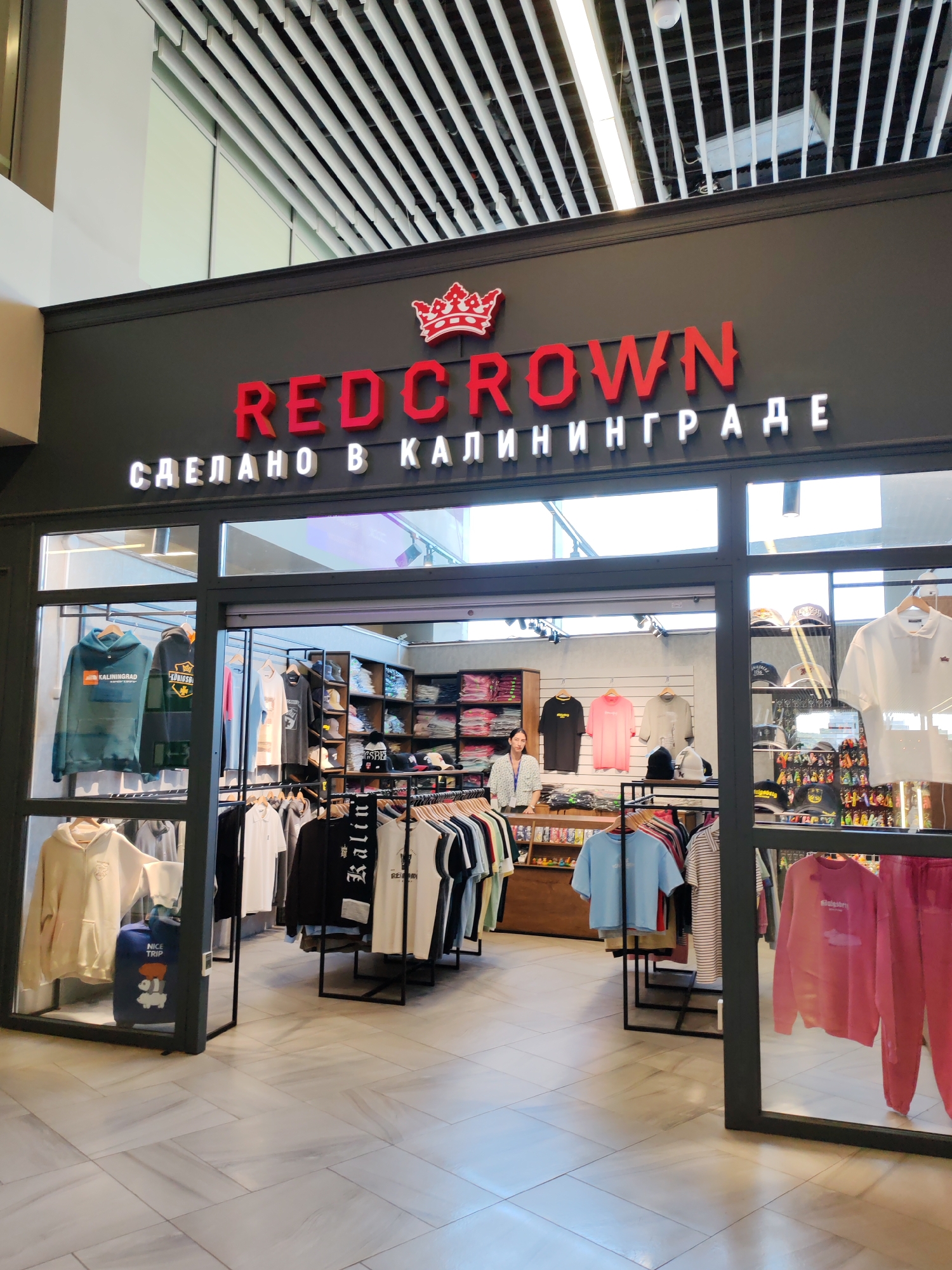 The image shows the storefront of a clothing store named "REDCROWN" with the text "СДЕЛАНО В КАЛИНИНГРАДЕ" (Made in Kaliningrad) below it. The store appears to be located within an indoor shopping mall, as indicated by the modern ceiling with parallel white beams and lighting fixtures. Through the storefront windows, racks of clothing, including hoodies, t-shirts, and hats, are visible. A woman is standing behind a counter inside the store. The floor is tiled with a light-colored, patterned material. The exterior of the store has a dark gray facade with the prominent red "REDCROWN" lettering and a red crown logo above it. The overall impression is that of a retail establishment selling casual wear. The mention of Kaliningrad in the store's signage suggests that the city of Khrabrovo, Russia, which is near Kaliningrad, might be the location of this store, or that the store specializes in products made in Kaliningrad.