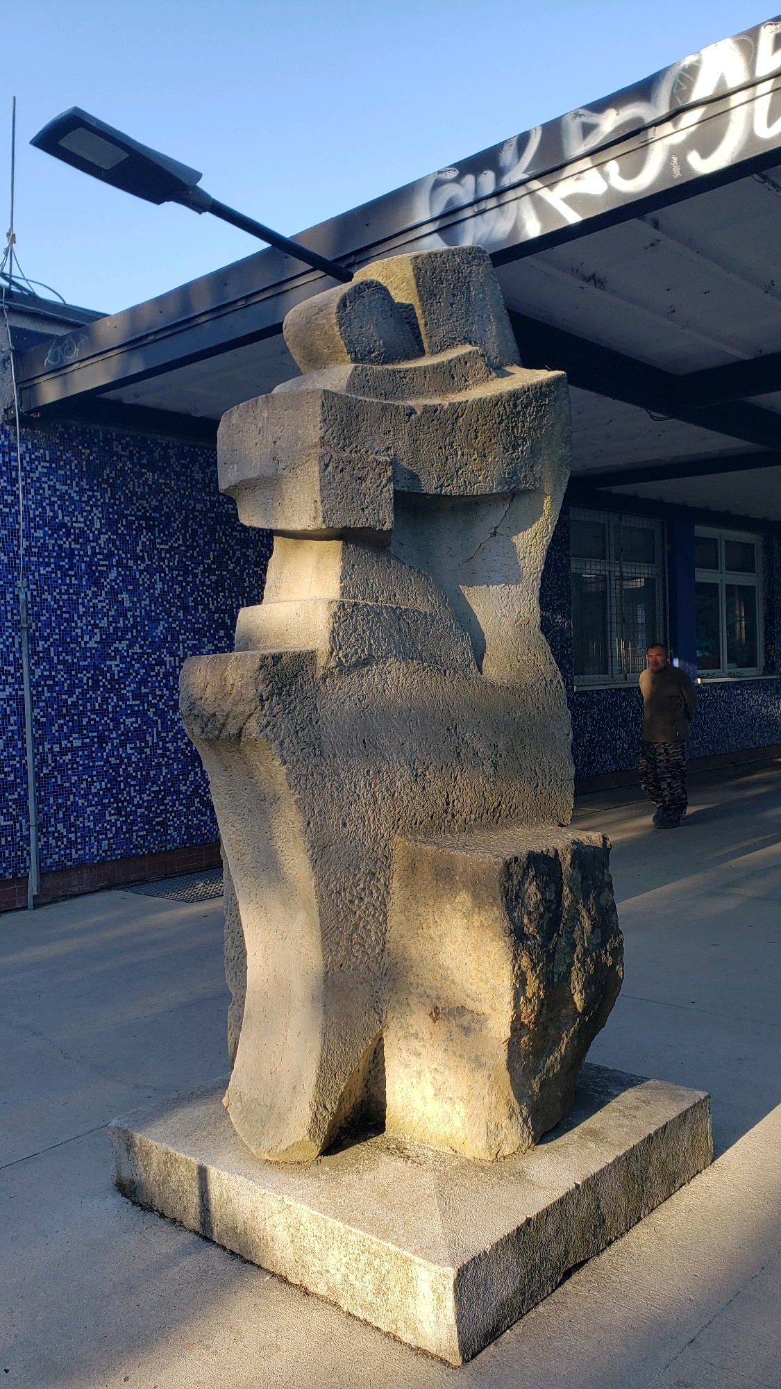 The image depicts a large, abstract concrete sculpture situated outdoors in front of a building. The sculpture has a rough, textured surface and is composed of interlocking geometric shapes. It is placed on a square concrete pedestal.
In the background, there is a building with a facade covered in small blue tiles. Above the tiled section, there is a dark overhang or roof structure. Graffiti is visible on the underside of this overhang. A street lamp is mounted on the overhang, extending over the area.
To the right of the sculpture, near the building, a person is standing. They are wearing camouflage-patterned pants and a dark-colored top. The person is looking towards the left, in the general direction of the sculpture. The ground is paved with concrete. Sunlight casts shadows on the ground and illuminates parts of the sculpture.
The location is identified as the area in front of the Primary School on Šmeralova Street in Prešov, Slovakia.
