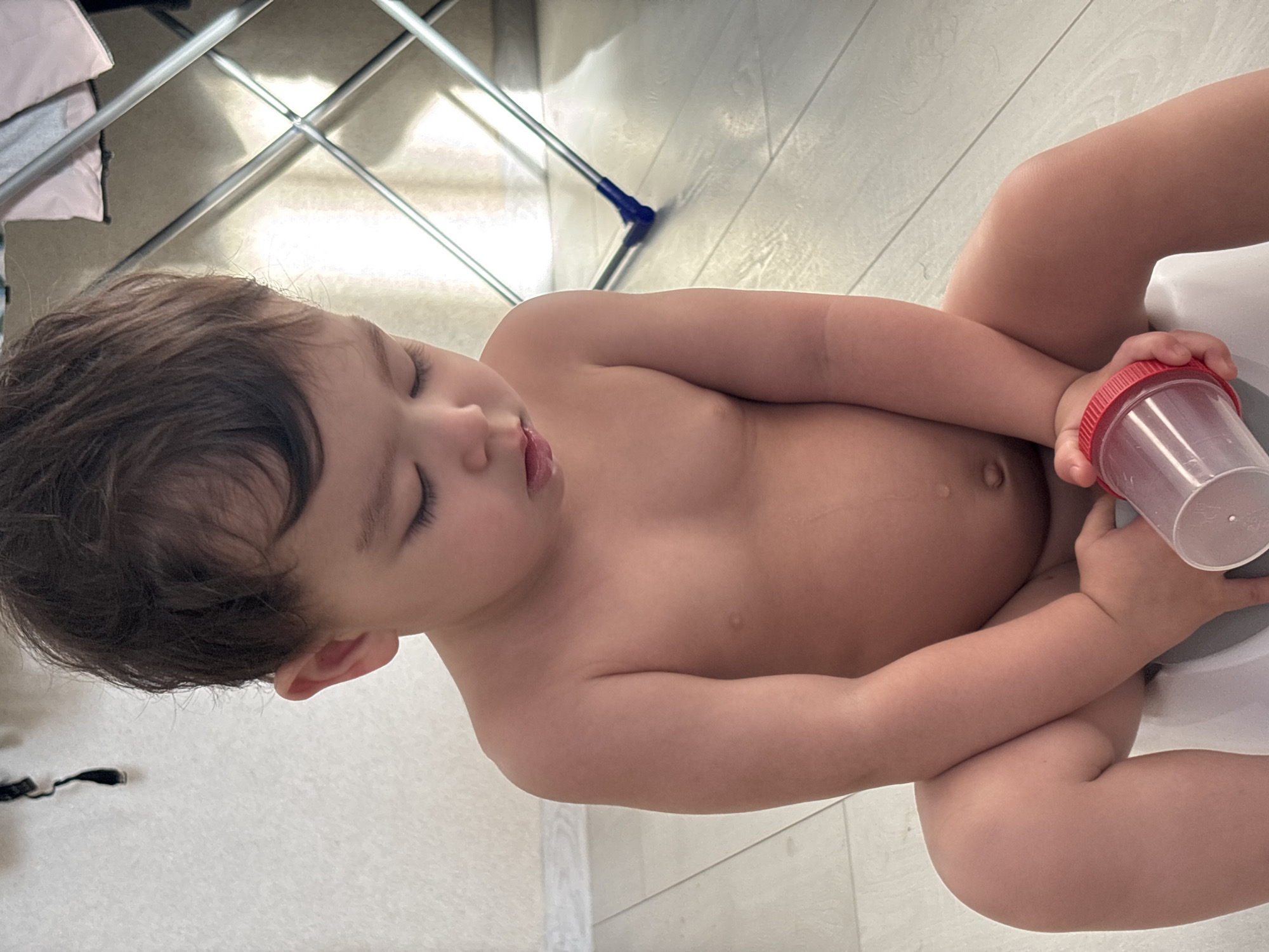 A partially clothed young child sits on a white potty chair, holding a clear plastic container with a red lid. The child appears to be sleeping or resting, with eyes closed. The location appears to be indoors, on a light-colored tiled floor. Behind the child, a metal clothes drying rack is visible. There is no information in the image to determine if the photograph was taken in the city of Chaikovsky, Russia.