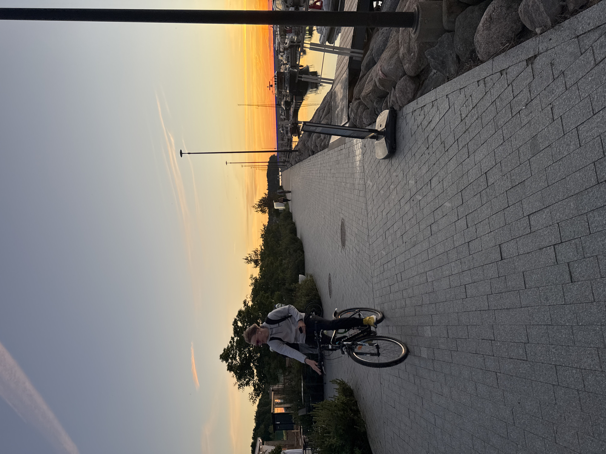 A young man with blond hair and glasses is riding a bicycle along a paved walkway. He is wearing a grey hoodie and dark pants. The walkway is bordered on one side by lush green bushes and trees, and on the other by a rocky shore with a harbor filled with boats in the distance. The sky is a gradient of soft blue and vibrant orange, indicating either sunrise or sunset. The scene appears to be in a coastal area of Tallinn, Estonia, likely at a marina or waterfront promenade. The lighting suggests it is late evening or early morning.