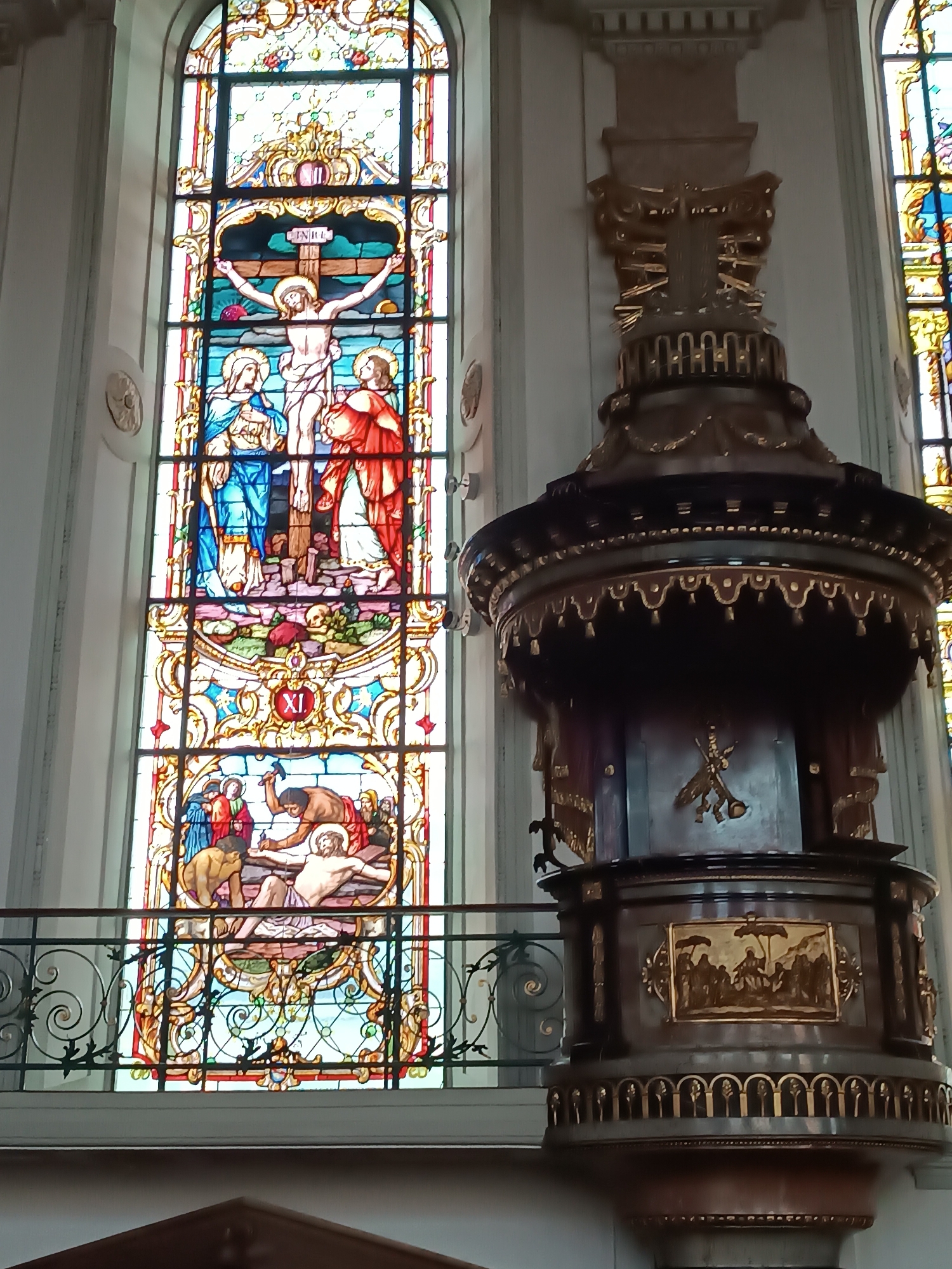 This is an interior photograph of a church, likely located in Appenzell, Switzerland, given the presence of stained glass windows depicting religious scenes. On the left side of the frame, a tall, arched stained glass window occupies a significant portion of the image. The window is divided into multiple panels, each illustrating a scene from the life of Christ. The top panels depict the crucifixion of Jesus on the cross, with figures of Mary and John at the base. Below this, a panel displays Roman numerals "XI" within a circular emblem, followed by another panel showing the entombment of Christ, with figures of mourners and individuals preparing his body. The stained glass is vibrant, with rich colors and intricate details.
To the right of the stained glass window is a tall, ornate wooden pulpit, characteristic of church architecture. The pulpit is dark wood with elaborate gold detailing, including decorative trim and religious iconography. It has a large, bell-shaped structure at the top, tapering down to a circular base. A detailed gold relief panel depicting a scene of people gathered under umbrellas is visible on the lower section of the pulpit.
A black wrought-iron railing runs horizontally across the lower part of the image, separating the viewing area from the stained glass window and pulpit. The background walls are a neutral color, suggesting a well-maintained interior. The lighting in the scene appears to be a mix of natural light filtering through the stained glass and interior lighting, casting soft shadows. The overall impression is one of solemnity and religious devotion.
