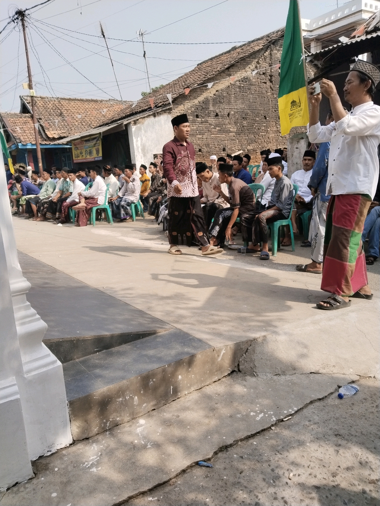 In the city of Serang, Indonesia, a group of men, predominantly wearing Islamic attire such as peci (caps) and sarongs, are gathered outdoors in what appears to be a residential or community area. The scene is characterized by a slightly elevated concrete ground with a low, dark-colored step leading up to a raised platform. A white pillar is visible on the left side of the frame.
In the foreground, a man in a white shirt and colorful checkered sarong is standing and holding a phone up, as if taking a picture or video. He is facing towards a group of other men seated on chairs. A green and yellow flag with Islamic calligraphy is positioned behind him. Another man in a maroon patterned shirt and sarong is standing and looking towards the right side of the frame.
The seated men are arranged in rows, facing forward, and appear to be listening or observing an event. They are dressed in various colors and patterns of shirts and sarongs, with many wearing peci. The background reveals a row of buildings with tiled roofs, some appearing to be simple residential structures, and a prominent brick wall. Electrical wires crisscross the sky above, suggesting an urban or semi-urban setting. The ground is cracked in places, and a plastic water bottle is visible on the ground. The overall impression is of a community gathering or religious event.