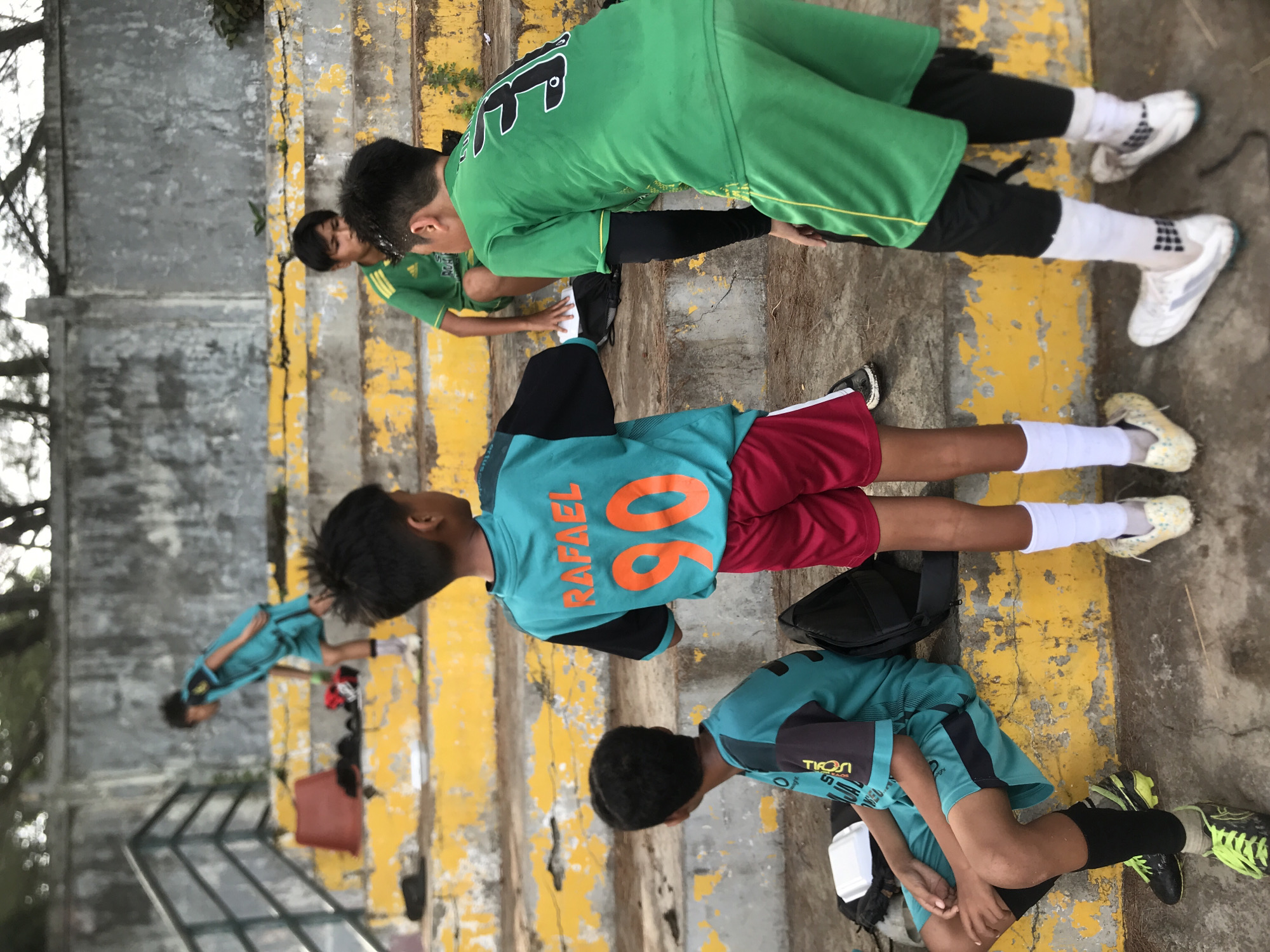 The image depicts several young individuals, likely a sports team, on concrete steps marked with yellow stripes. The team appears to be stretching or warming up. Most are wearing soccer attire, with different jerseys and shorts in green, maroon, and blue-green color schemes. At least one jersey has the name "RAFAEL" and the number "06" visible. The individuals are positioned at different levels of the steps. The surroundings are a mix of concrete and greenery. The city of Lhokseumawe, Indonesia, is not explicitly confirmed but can be inferred from the general context of Southeast Asian youth playing soccer.