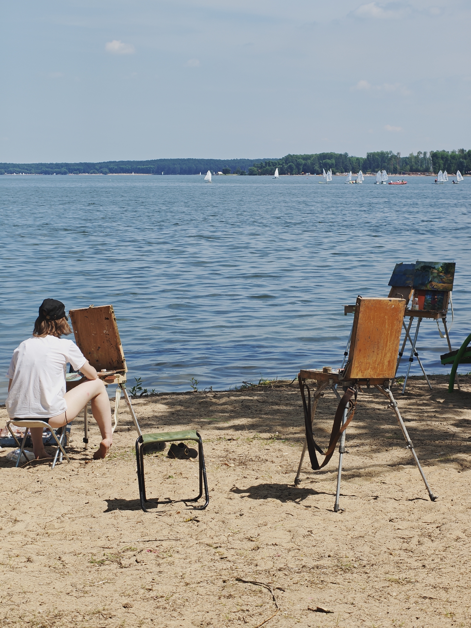 A person sits on a small stool on a sandy beach, facing a lake and an easel. The person is wearing a white t-shirt and black shorts, with a black baseball cap. Their legs are crossed, and their bare feet are visible. They are engaged in painting, as evidenced by the easel and what appears to be a palette and brushes in a small box on the easel.
In the background, the lake stretches out to the horizon, dotted with numerous sailboats. A treeline borders the far side of the lake, suggesting a natural setting. The sky is a clear blue with a few scattered clouds. The sun appears to be high in the sky, indicating it is likely midday.
Another easel stands to the right of the person, with a partially completed painting on it. This easel is also positioned on the sand. A smaller, green folding stool is placed between the two easels. The overall scene suggests a peaceful, artistic outing in a scenic location. The sand is lightly textured with small pebbles and dry grass. The water is calm, with gentle ripples. The lighting suggests a bright, sunny day. The location is identified as Ждановіцкі сельскі Савет, Belarus. No visible text is present in the image.