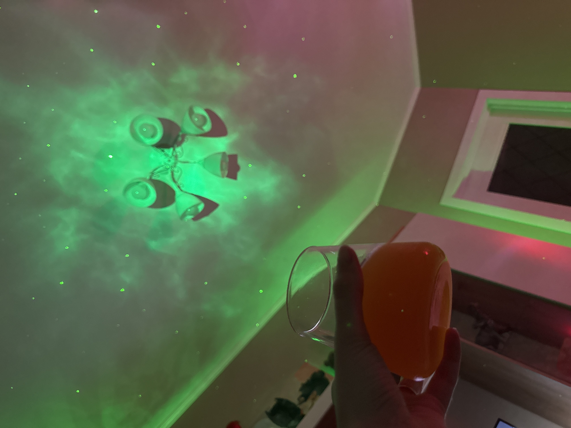 A low-angle shot focuses on a hand holding a glass filled with orange liquid. The ceiling, illuminated with a green light and small, point-like green specks resembling stars, suggests a domestic interior. A light fixture with a cluster of bulb-like forms is also on the ceiling. A window is in the background, and hints of furniture, possibly a shelf or cabinet, are visible behind the hand. The lighting and focus give the scene an otherworldly feel. Location: Unknown, Unknown.