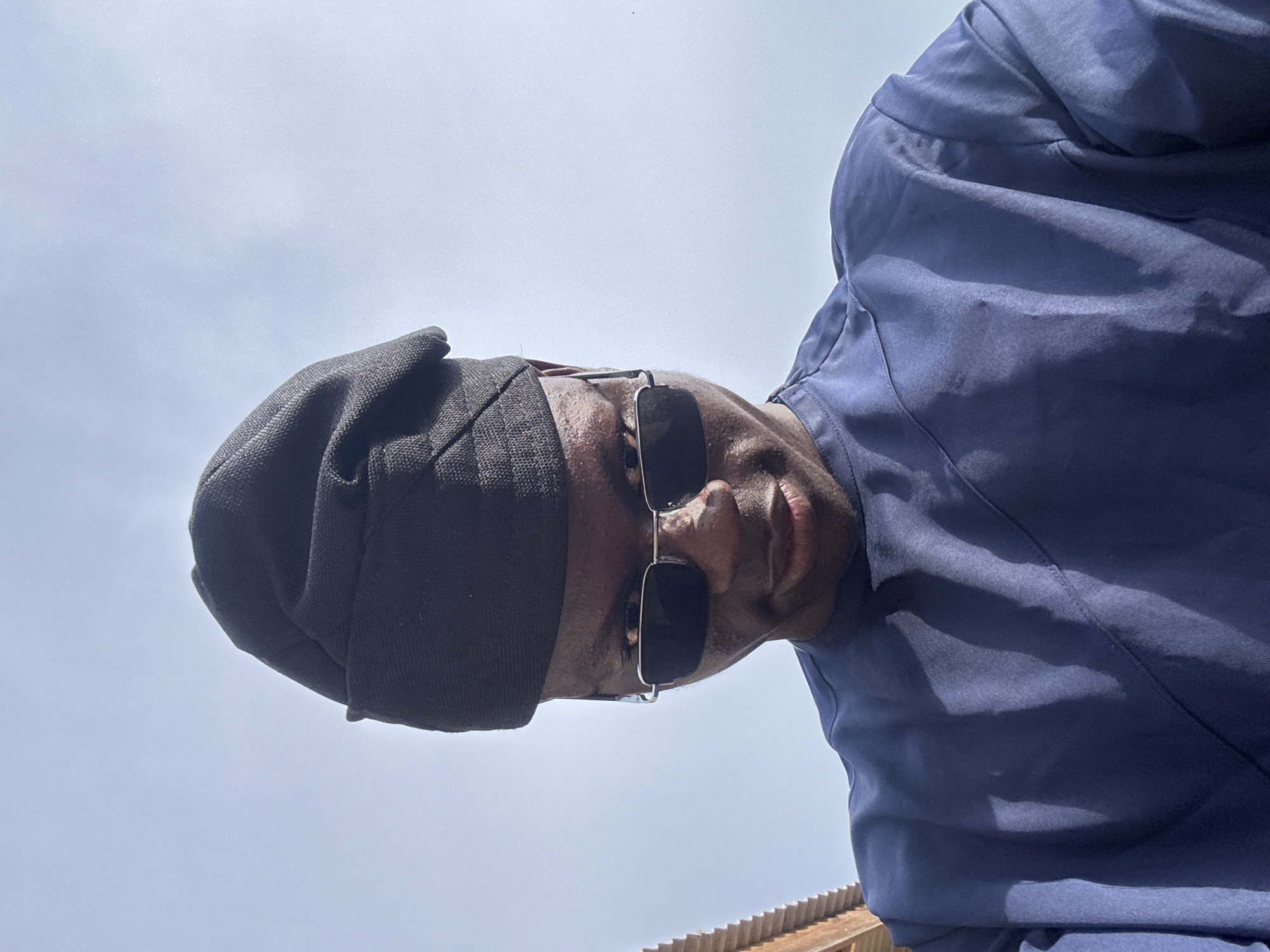 A low-angle shot of a person with dark skin, wearing a dark, woven hat and sunglasses with silver frames. They are also wearing a blue shirt. The background consists of a light sky and what appears to be a corrugated metal roof, possibly in the city of Badagry, Nigeria, though this cannot be definitively confirmed from the image alone.